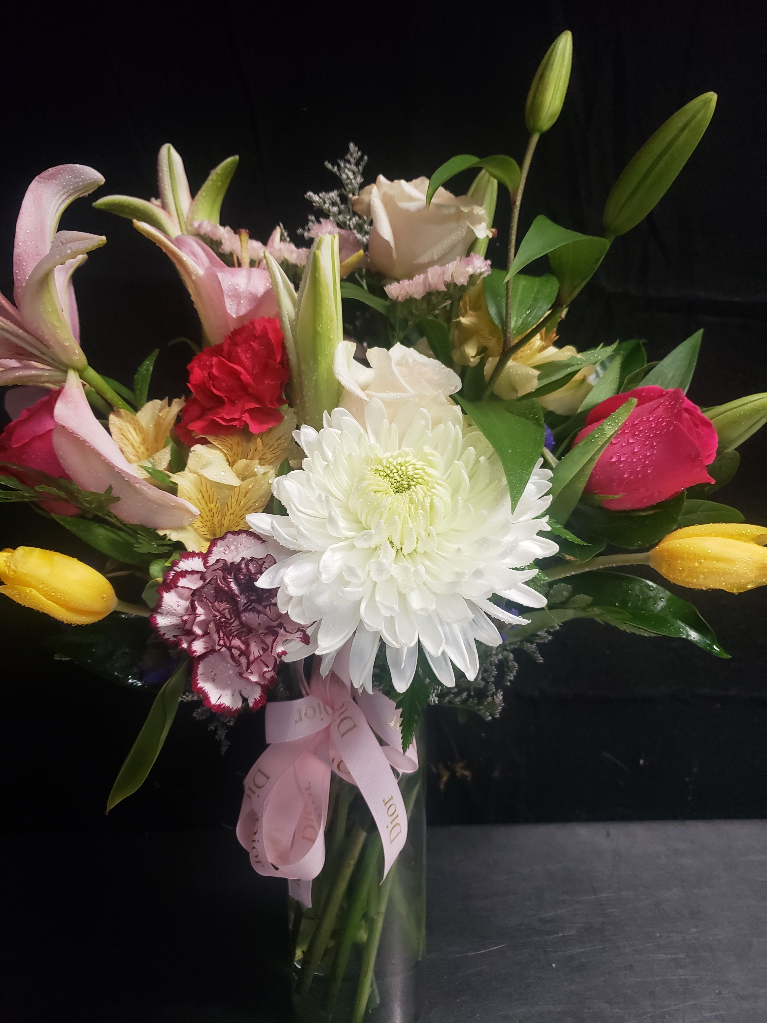 SURPRISED BOUQUET - A lush cluster of white lilies and a large white chrysanthemum anchors this mixed bouquet, the first impression bright and refined - perfect as a birthday bouquet, thank-you arrangement, or romantic gift. Pink and red roses mingle with pink and white lilies, carnations, and alstroemeria for a layered, full composition that reads both celebratory and intimate; yellow accents lift the palette so the recipient feels genuinely cheered and noticed.  Arranged upright in a clear glass vase and finished with a pink ribbon, the design pairs open blooms with unopened lily buds for height and texture, framed by broad green leaves for contrast. The colors-soft pinks and whites warmed by red and yellow-create a versatile gift that suits many occasions. Sized to sit on a table or mantle, this vase arrangement arrives ready to display; change the water every few days and trim stems to keep blooms fresh longer.