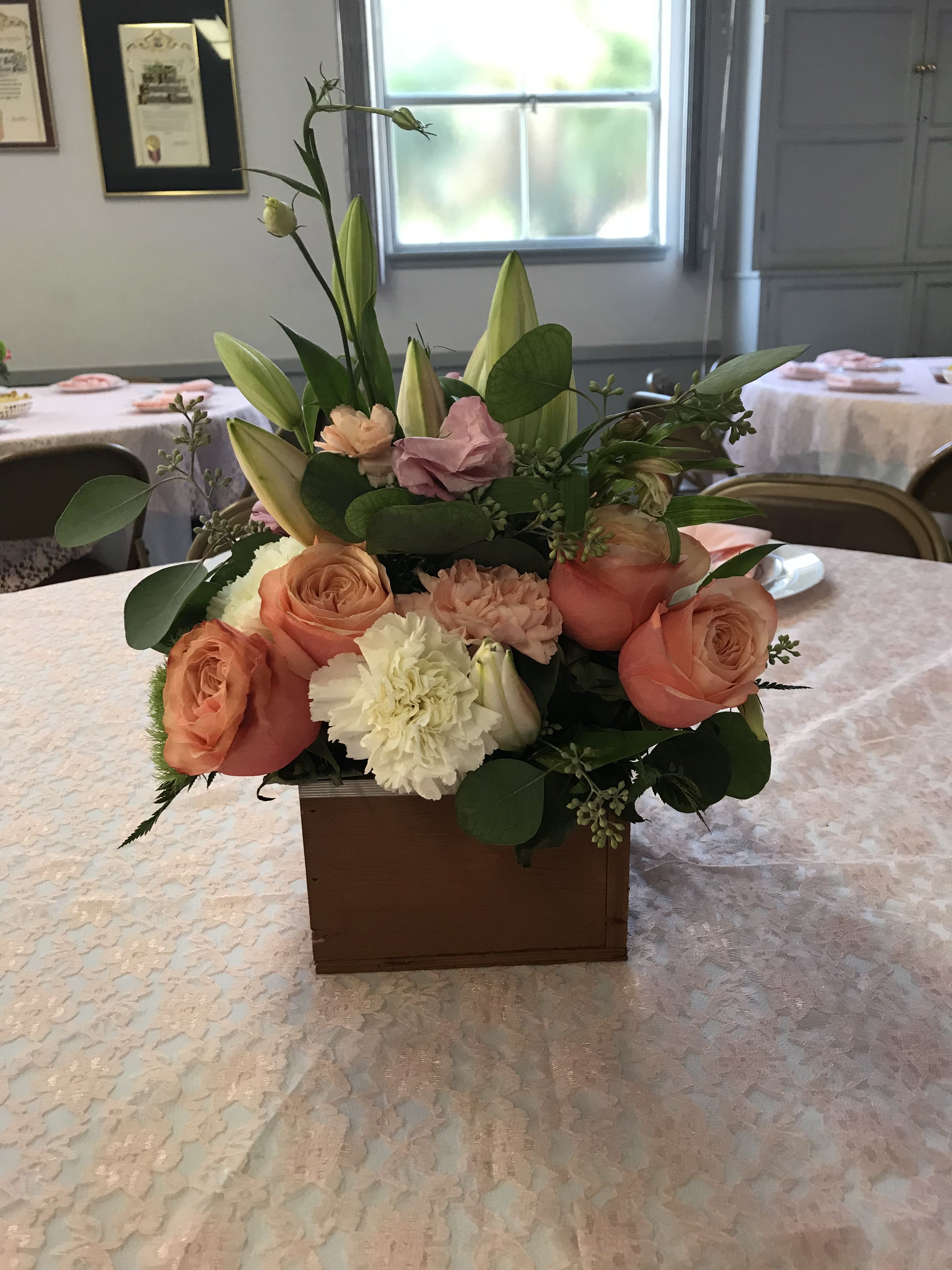 CENTROS DE MESA - A low, romantic centerpiece in a rectangular wooden box blends peach roses, white lily buds, and blush-and-white carnations into a soft, layered display perfect for weddings, bridal showers, or elegant receptions. The taller lily buds lift the composition above the compact mass of rounded blooms, so the person you're honoring will feel gently celebrated without the flowers blocking conversation at their table.  For taller table settings, choose a clear cylindrical vase filled with a dense dome of white hydrangeas accented by pink and orange roses to create an airy, formal focal point. These mixed-color arrangements work across round banquet tables dressed in white linens with pink chair sashes and gold chargers; they read beautifully in a coordinated room of repeated centerpieces and tall clear glass vases.  Whether you want low wooden-box pieces for intimate guest tables or tall vase arrangements to give a ballroom a cohesive, polished look, the palette of peach, pink, white, cream and green brings a timeless, romantic mood. We recommend grouping low and tall centerpieces for visual rhythm; standard sizing fits typical banquet tables and these designs arrive ready to place so your event tables look unified from the first guest to the last dance.