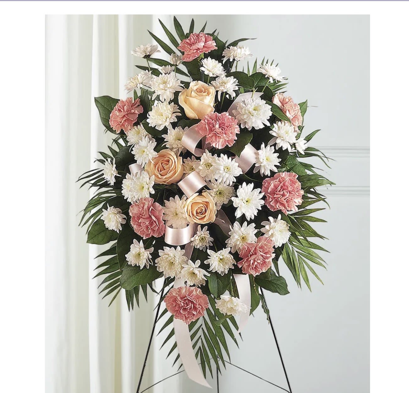 FUNERAL, SYMPHATY - A large standing sympathy spray that reads as quietly elegant and deeply respectful, perfect for a funeral or memorial service. Soft pinks, peaches and whites-carnations, roses, lilies, chrysanthemums and small daisy-like blooms-are arranged in a full, rounded profile so the person you're honoring feels remembered with grace. The pastel palette (with occasional purple or blue accents) and looping pink satin ribbon create a gentle visual that comforts mourners and suits a formal ceremony.  These substantial sprays and wreaths arrive mounted on wooden or standing easels and are finished with white ribbon banners, a small card pocket and tasteful ribbon accents. Expect dense groupings of blooms framed by dark green foliage and palm fronds for height and structure; some designs feature vivid blue roses set against a white border for a striking, traditional memorial statement. Order size options and personalization for ribbon text are available-please let us know the memorial wording for the ribbon and the name to include on the card so we can prepare the arrangement for the service.