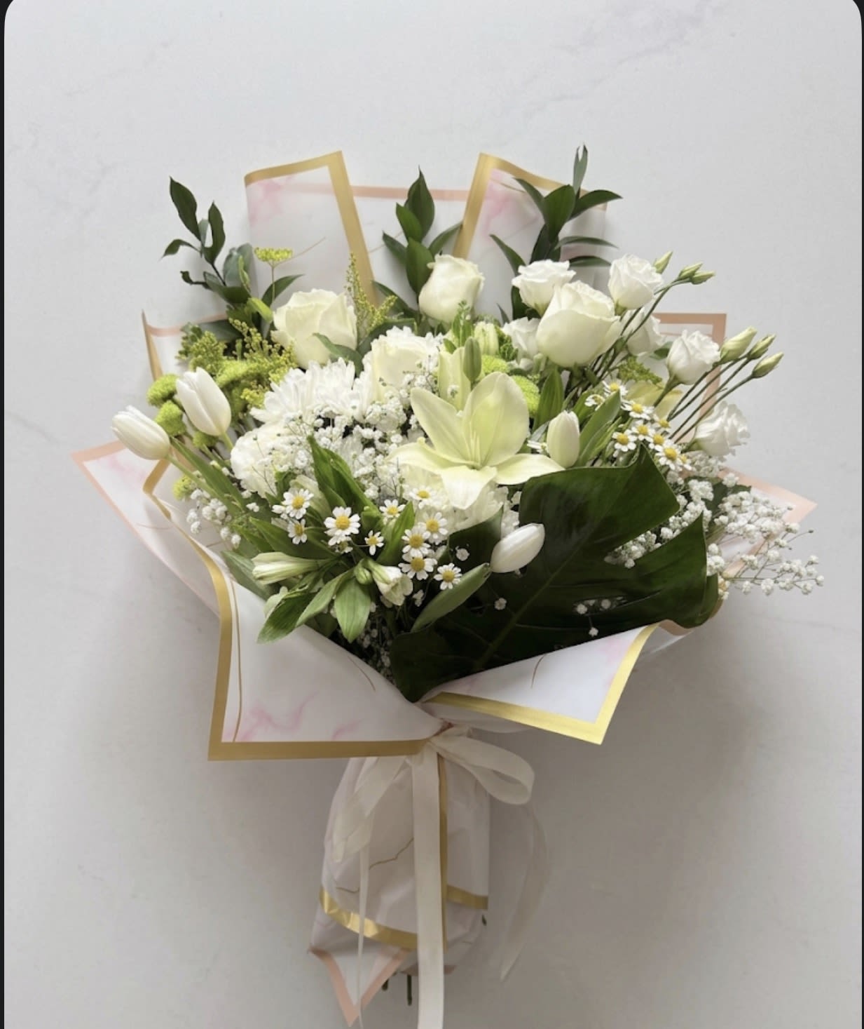 SYMPHATY - Soft white lilies and roses take center stage in this sympathy flower arrangement, designed to offer calm and comfort for moments of remembrance. Framed by pale blue hydrangea and gentle purple accents, this arrangement works beautifully as a sympathy or memorial bouquet while also serving as a refined choice for a thoughtful get-well or condolence gift-the kind of floral expression that lets the person you're thinking of feel seen and gently supported.  The arrangement combines white roses, a prominent pale lily, daisies and alstroemeria with textured filler blooms and deep green foliage for contrast. One presentation is hand-tied in translucent paper with gold edging and finished with a satin ribbon for a soft, gift-ready look; another is arranged full and rounded in a clear glass vase with layered blooms and a few long, arching leaves that add movement. The palette of white, green, pale blue and touches of purple reads fresh and understated, with a refined, polished silhouette that complements both home display and formal settings.  Ideal for sending sympathy, expressing condolences, or bringing a quietly comforting presence to a room, this arrangement arrives ready to display (vase option) or present (hand-tied option). Care is simple: keep flowers in fresh water, trim stems and refresh water every couple of days to help blooms last and maintain their serene appearance.