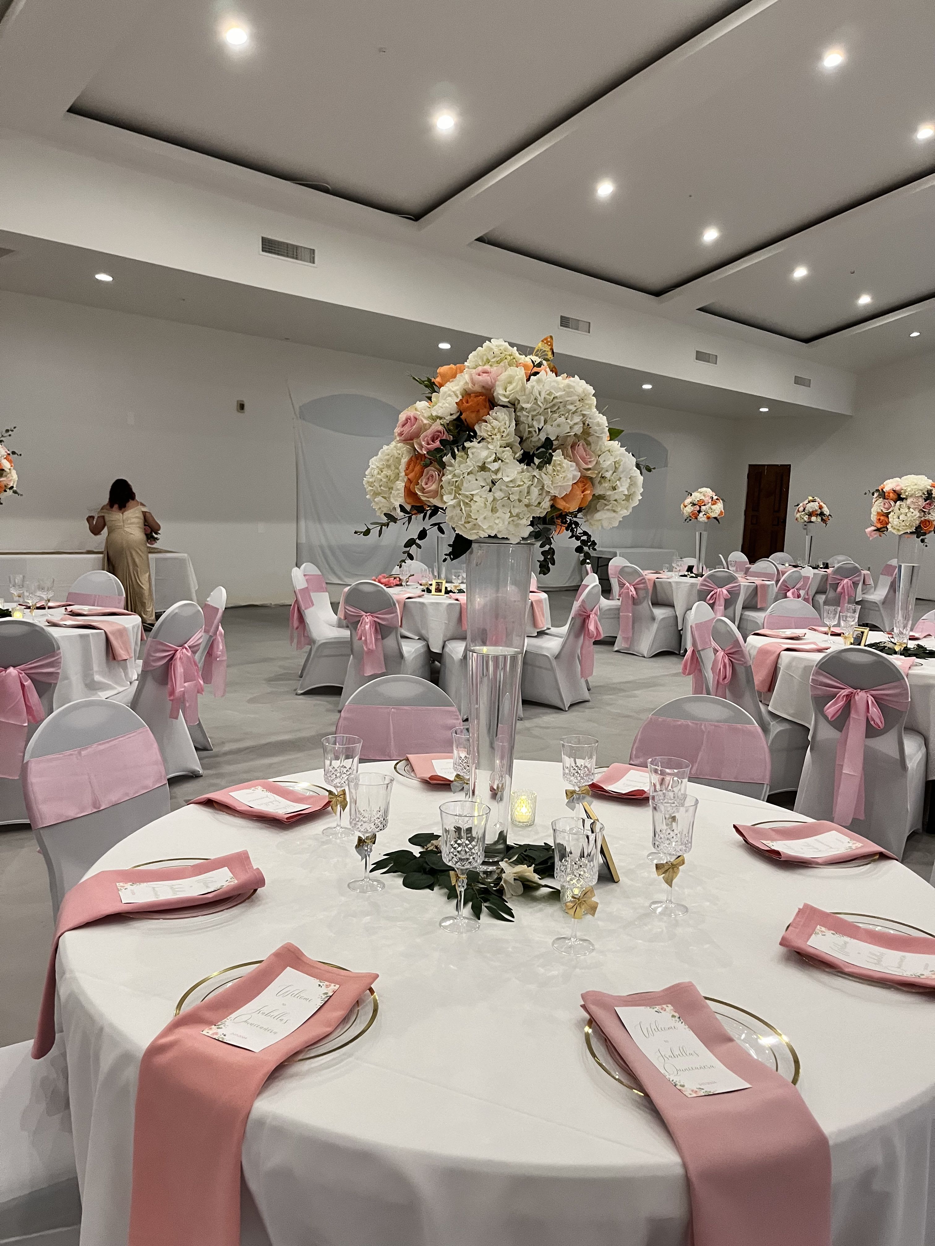 EVENTO FIESTAS - A tall banquet centerpiece that reads like a pastel celebration-this event flower arrangement is perfect for a wedding reception or formal party. The rounded mass of white hydrangeas gives an airy, cloud-like backdrop while pink and orange roses peek through to add romantic warmth; the look says "celebration" without shouting, making it ideal for anniversaries, bridal showers, or upscale birthday dinners.  Set in a clear cylindrical glass vase, the arrangement includes a few darker greenery accents that trail just beneath the bloom mass for a touch of natural contrast. The palette of white, soft pink, and muted orange reads beautifully against coordinated table linens, white chair covers, and pink chair sashes shown in the room-imagery that helps recipients picture the full event styling. Designed to sit tall on banquet tables, it creates presence without blocking conversation.  For a bride, guest of honor, or the person you want to make feel truly seen, this centerpiece frames the moment with elegance and charm. Order details: delivered in the clear glass vase shown and sized for banquet tables-simple care is keeping stems fresh and replacing water as needed to maintain that airy, just-arranged look through the event.