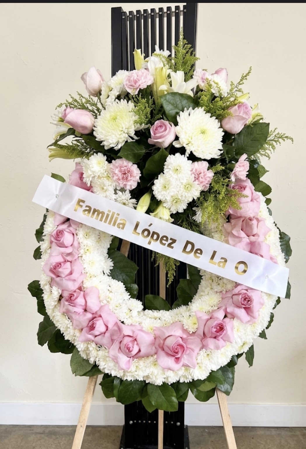 FUNERAL - A large standing funeral wreath that offers a soft, respectful tribute for sympathy or memorial services. The circular arrangement of pink roses, white chrysanthemums, and white lilies creates a gentle, formal expression of condolence the person you're honoring will receive as a quiet, heartfelt gesture. The white memorial ribbon with gold lettering across the front reinforces the tribute and reads as a dignified statement for a service or graveside ceremony.  Flowers are densely packed in a classic round design with dark green foliage framing the outer edge and filling the center for a full, finished silhouette. Pink roses add warmth and tenderness, while white chrysanthemums and lilies lend purity and peace; mounted on an easel and finished with the memorial ribbon, it arrives ready to display. Sized for visibility at a service, this arrangement is designed for immediate presentation-no assembly required-and communicates sympathy in a gentle, traditional way.