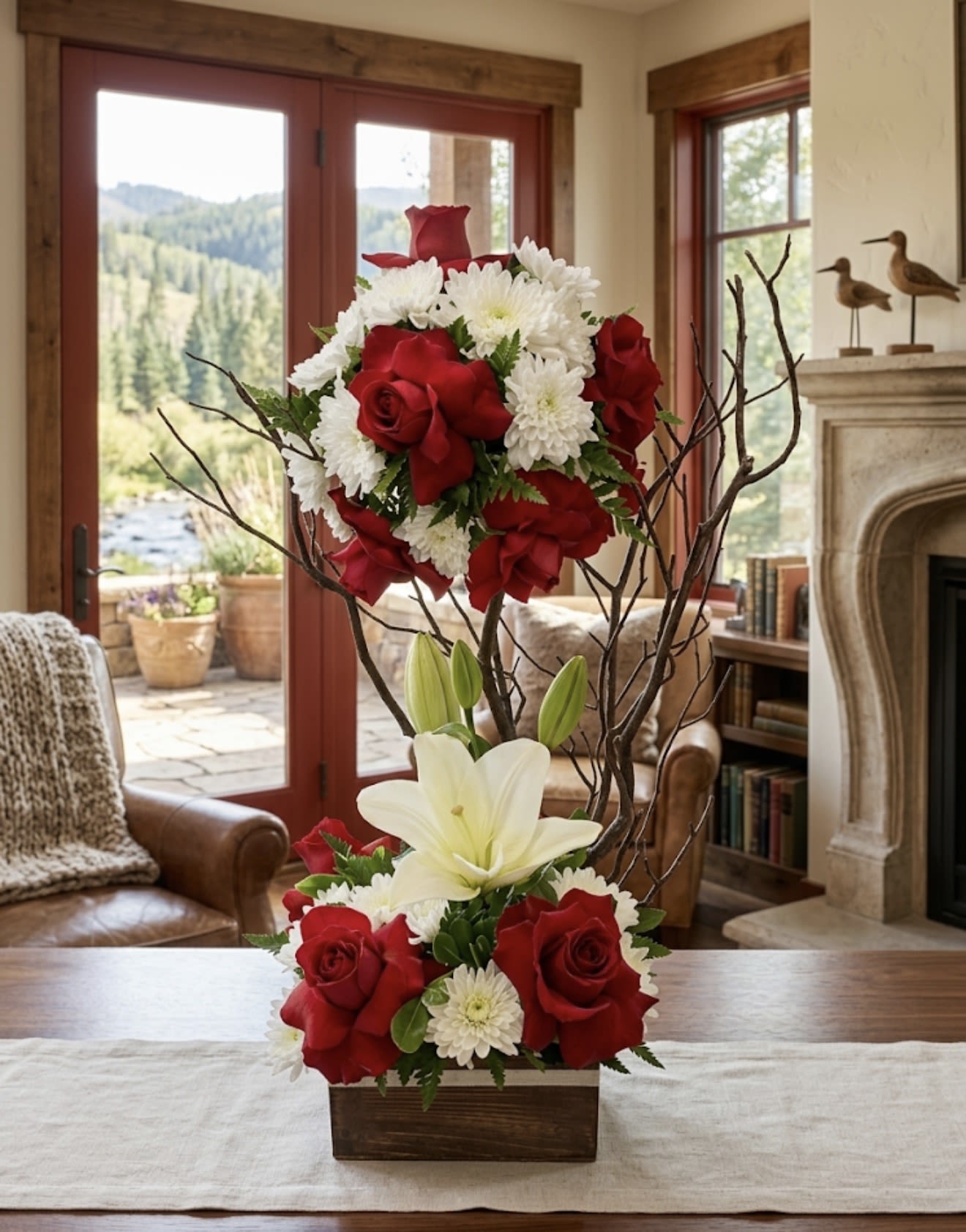 RAMA DE MANZANITO  - A sculptural, upright floral arrangement that reads formal and romantic-perfect as a birthday bouquet, anniversary gift, or elegant sympathy arrangement. Two clustered sections of red roses and white chrysanthemums are joined by a prominent white lily and tall, bare decorative branches, giving the piece a dramatic vertical presence that will make the person you're thinking of feel both cherished and noticed.  Set in a rustic wooden box, the red-and-white palette is bright and classic, balanced by green foliage and the warm texture of the wood and branches. The lilies add a soft fragrance and focal contrast to the velvety roses and full, cushiony chrysanthemums, creating visual depth from tabletop to eye level. Its upright profile suits an entryway, dining table centerpiece, or office reception spot-order with confidence for a meaningful, polished statement that lasts through the occasion.