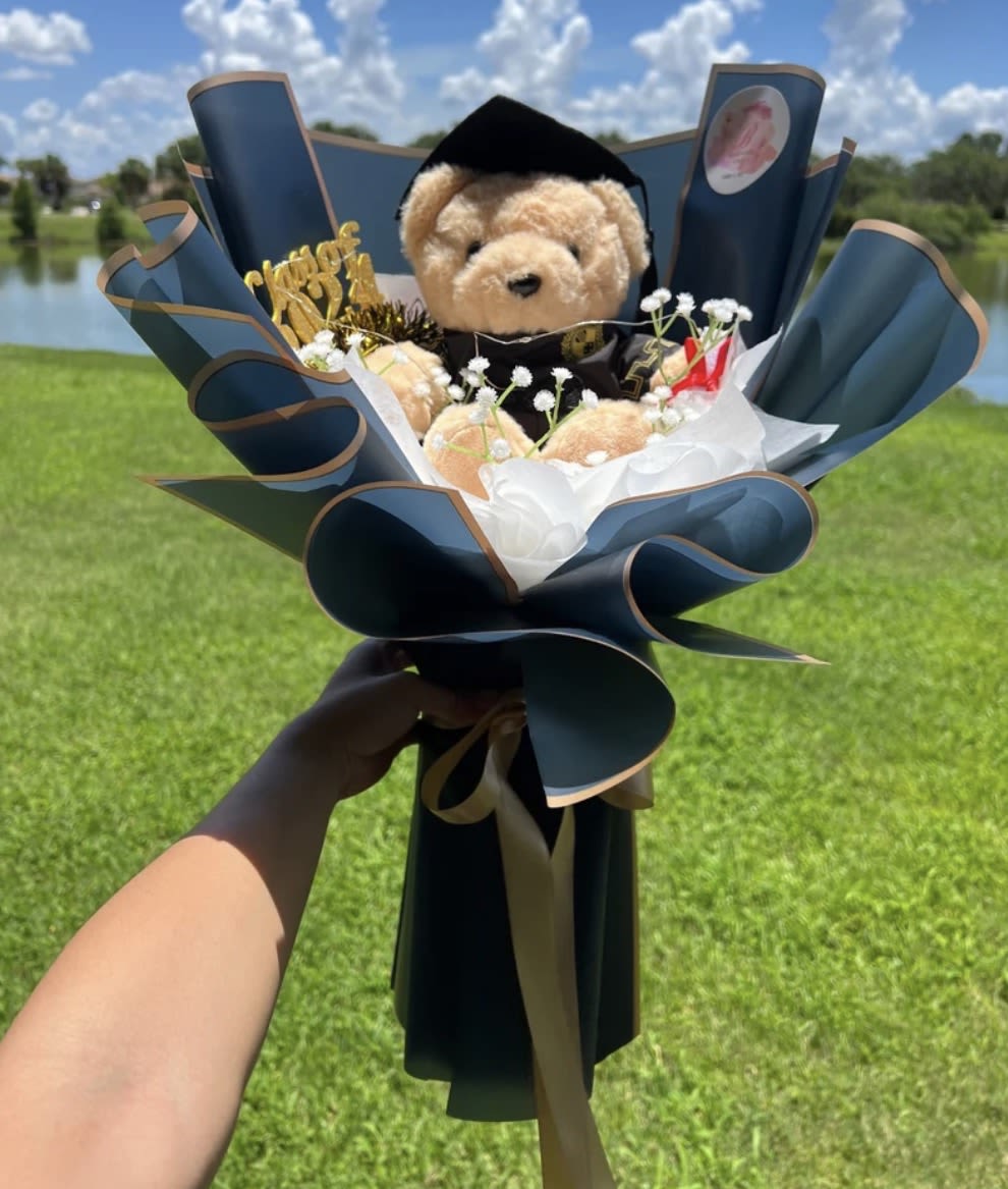 GRADUATES - A plush teddy bear peeks out from a dramatic dark blue paper wrap edged in tan-this graduates flower arrangement is a playful, celebratory gift perfect for a graduation, congratulations, or birthday bouquet. The bold contrast of deep blue, black accents, soft pink touches, and bright white filler creates an eye-catching presentation that feels festive and personal-the kind of gift that makes the person you're thinking of feel noticed and celebrated.  Rather than a traditional vase of blooms, this arrangement centers on the cuddly keepsake and is finished with a gold topper, ribbon, and decorative paper that give it a party-ready polish. Small white filler flowers surround the bear for a delicate, airy texture against the rich wrap. Sized for hand-held gifting, it arrives ready to present and makes an immediate, joyful impression for graduates, achievers, or anyone marking a milestone.