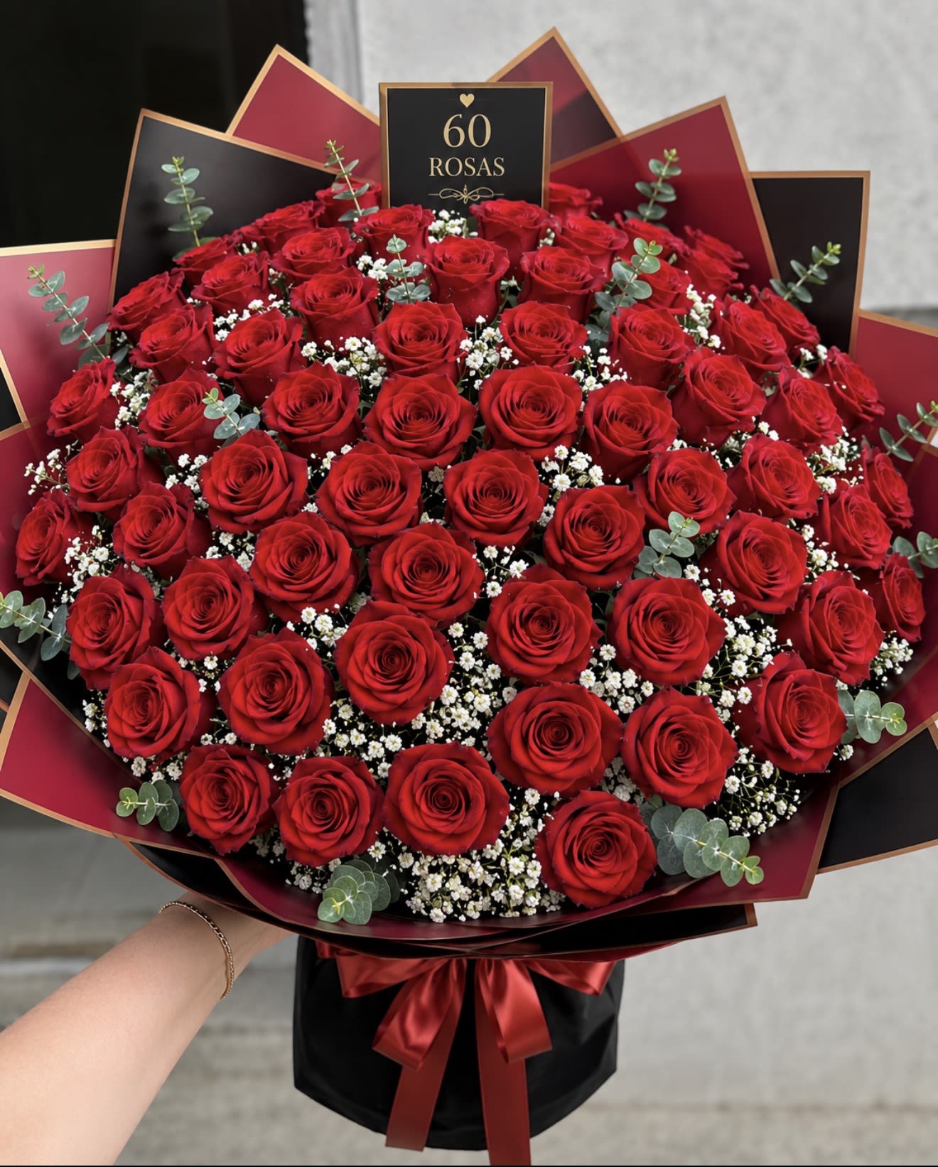 BUCHONES 60 ROSAS - A dramatic dome of sixty red roses designed for milestone celebrations and anniversary bouquets that make the recipient feel deeply celebrated. Rich red roses form a dense, rounded mass while tiny white filler blooms and soft eucalyptus sprigs peek through to add texture and a cool green contrast-perfect for saying "60" or marking a major anniversary with bold, romantic flair.  Wrapped in layered black and burgundy decorative paper and tied with a red satin ribbon, this arrangement reads as elegant and theatrical-ideal for anniversaries, milestone birthdays, or any moment meant to feel unforgettable. The included card reads "60 ROSAS," and the bouquet comes finished with a coordinating ribbon so it arrives ready to present; to keep roses lasting longer, place in fresh water and trim stems at an angle before displaying.