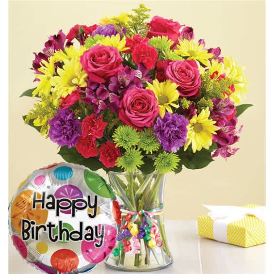 Mixed bouquet of pink roses, yellow daisies, and purple flowers in a glass vase with a birthday balloon.