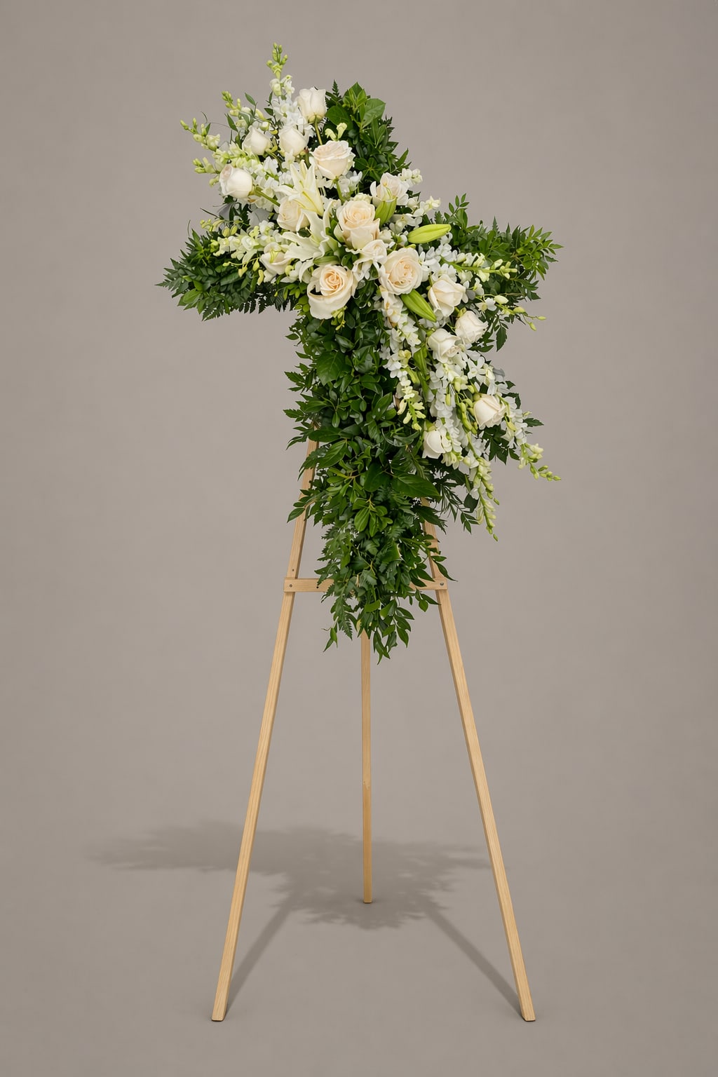 Ivory Peace Cross - A tall, reverent standing tribute sculpted into a classic cross silhouette, this sympathy flower arrangement is made for moments of remembrance and funeral service display. The Ivory Peace Cross uses white roses and white lilies at the focal points to create a serene, comforting presence that communicates sympathy and respect.  Dense green foliage and small white filler flowers complete the cross form, with blossoms concentrated across the top and a trailing vertical section to emphasize the shape. Designed for display on a wooden easel, the arrangement reads formal and dignified from a distance while offering gentle texture and purity up close.  Perfect for honoring a loved one or expressing condolences to a grieving family, this piece delivers a calm, reverent message that helps make a memorial service feel thoughtful and composed. Please indicate delivery date and service details when ordering so we can ensure timely placement on the day of the funeral.