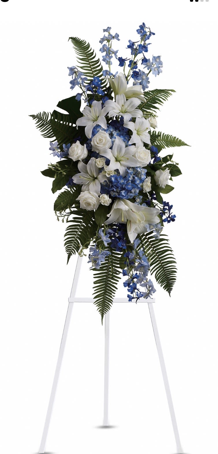 Ocean Breeze Spray - Tall white lilies open above clusters of blue hydrangea and sky-blue delphinium in this sympathy flower arrangement, creating the calm, formal presence of a standing spray for a funeral or memorial service. The white roses and layered fern fronds lend a gentle, graceful silhouette on a white easel stand, so the person you're honoring will be remembered with a serene, dignified display.  Crisp white, cool blues, and fresh greenery combine for a balanced mix of airy stems and dense blooms - lilies and roses provide focal blossoms while delphinium spikes and hydrangea clusters fill the composition with soothing color and texture. The upright, cascading shape reads well from a distance in a chapel or reception area and is designed to convey sympathy, respect, and peaceful reflection.  Appropriate for a funeral service or as a heartfelt tribute, this spray makes a thoughtful expression of condolence that helps loved ones feel seen and comforted. The arrangement arrives ready to display on the included white easel; please let us know any delivery details or service times so we can ensure it is placed with care.