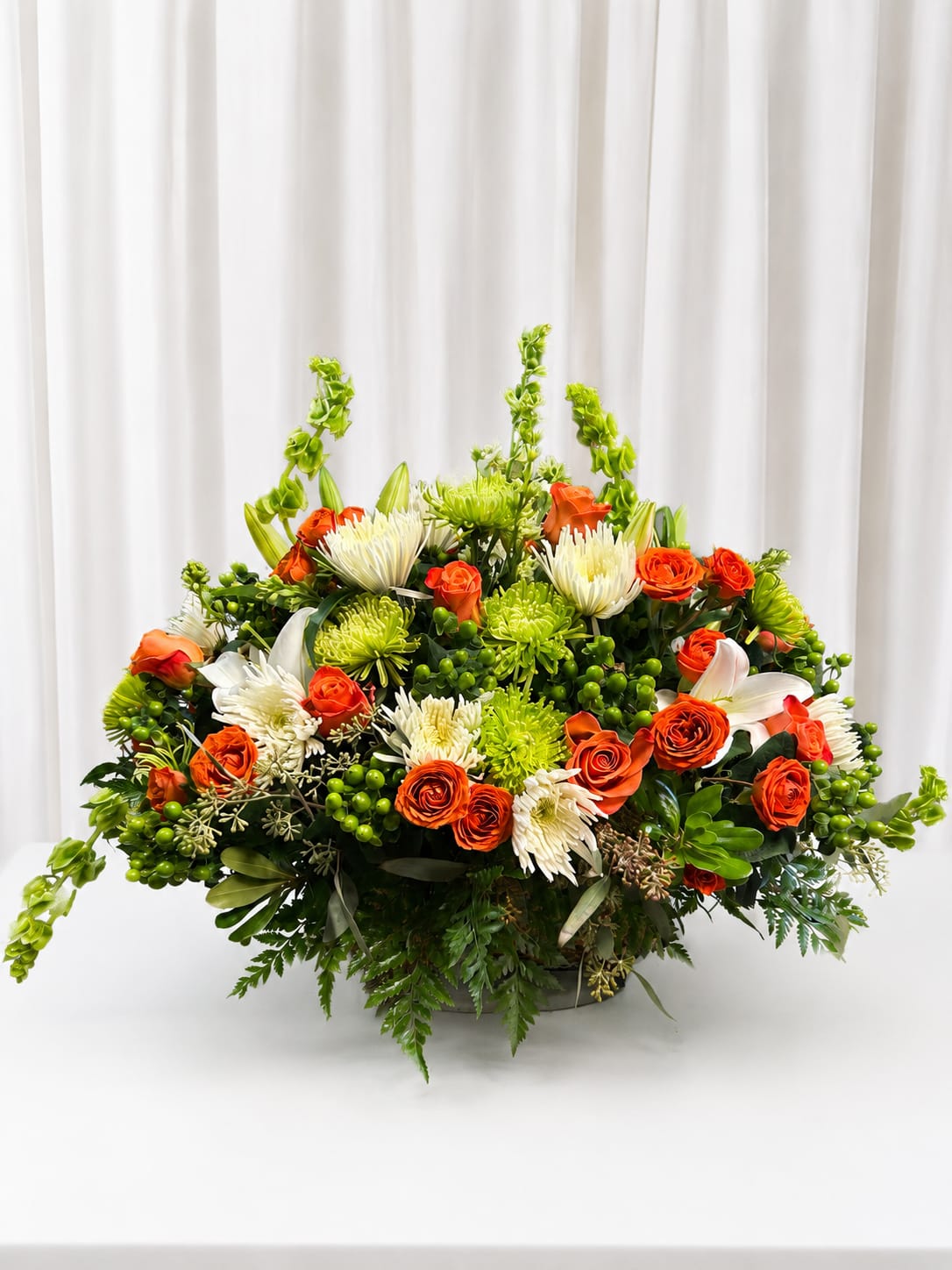 Clemers River glory - A bold, celebratory centerpiece that catches the eye with a warm orange-and-white palette-this large flower arrangement is perfect as a wedding centerpiece, anniversary display, or festive event accent. The rounded, low design lets the person you're honoring see and be seen, making it an effortless way to make someone feel celebrated and truly noticed.  Dense clusters of orange roses mingle with white spider chrysanthemums and open white lilies for a mix of soft petals and dramatic form. Bright green accent blooms and small berry-like clusters add lively texture and vivid contrast, while abundant foliage creates a lush, spilling shape from its low container. The composition reads formal yet energetic, offering both visual impact and a pleasing table-friendly profile.  Ideal for a reception table, entry console, or a special dinner, this arrangement conveys warmth and joyful sophistication. Because it sits low, it's easy to pair with place settings without blocking conversation; keep flowers fresh by changing the water every two days and trimming stems slightly when re-cutting. Order early for event delivery or pick-up to ensure this full, vibrant centerpiece arrives ready to impress.   If any design elements are of major importance to your order, please include them in the florist instructions at checkout or contact us to ensure availability.