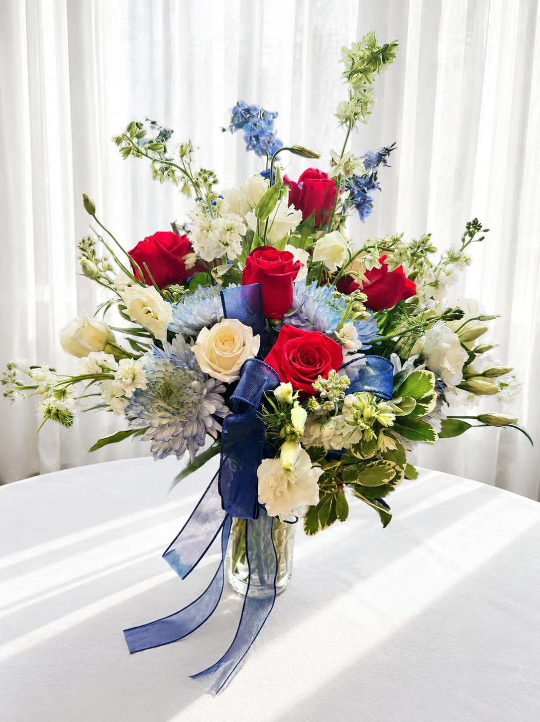 Stars and stripes - Vivid red roses and creamy white roses mingle with pale blue chrysanthemum-like blooms in a garden-style arrangement that reads instantly festive and refined - perfect as a patriotic centerpiece or a celebratory birthday or anniversary bouquet. Tall white and blue accent stems lift the composition into a layered, airy silhouette so the person you're thinking of will feel both noticed and celebrated the moment they see it.  This flower arrangement features roses, chrysanthemums, alstroemeria, and complementary seasonal blooms in a red, white, blue, and green palette set in a clear glass vase. A long blue ribbon tied around the vase adds a formal, polished touch that enhances the bright, balanced color story. The loose, natural grouping gives soft texture and movement while the taller accents provide height and presence for tabletops or entryway displays.  Ideal for Fourth of July gatherings, congratulations, or simply to brighten someone's day, this bouquet sends a clear message of joy and appreciation. Order with standard sizing for a graceful display - stems arrive arranged and ready to enjoy; refresh water every two days and recut stems as needed to keep blooms looking their best.   If any design elements are of major importance to your order, please include them in the florist instructions at checkout or contact us to ensure availability.