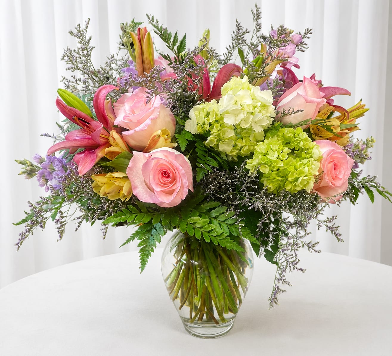 Sissy blooms - Bright, cheerful pink roses and pink lilies take center stage in this birthday bouquet-style arrangement, paired with lime-green hydrangeas for a fresh, springtime pop-perfect for birthdays, thank-you gifts, or simply to brighten someone's day. The mix of colors and textures creates a joyful, celebratory look that will make the person you're thinking of feel seen and cherished.  This rounded, abundant arrangement is arranged in a clear glass vase and includes yellow‑orange alstroemeria, fine purple filler flowers, and airy greenery that gives the design a slightly wild garden feel. The combination of soft romantic pinks, vibrant green, and playful yellow accents offers visual contrast and a lively scent profile from the roses and lilies.  Ideal for a friend, mom, or colleague who loves bright, garden-fresh bouquets, Sissy blooms delivers an easy, happy message of appreciation or celebration. It arrives ready to display in its clear vase-simply trim stems and refresh the water every few days to extend bloom life.   If any design elements are of major importance to your order, please include them in the florist instructions at checkout or contact us to ensure availability.