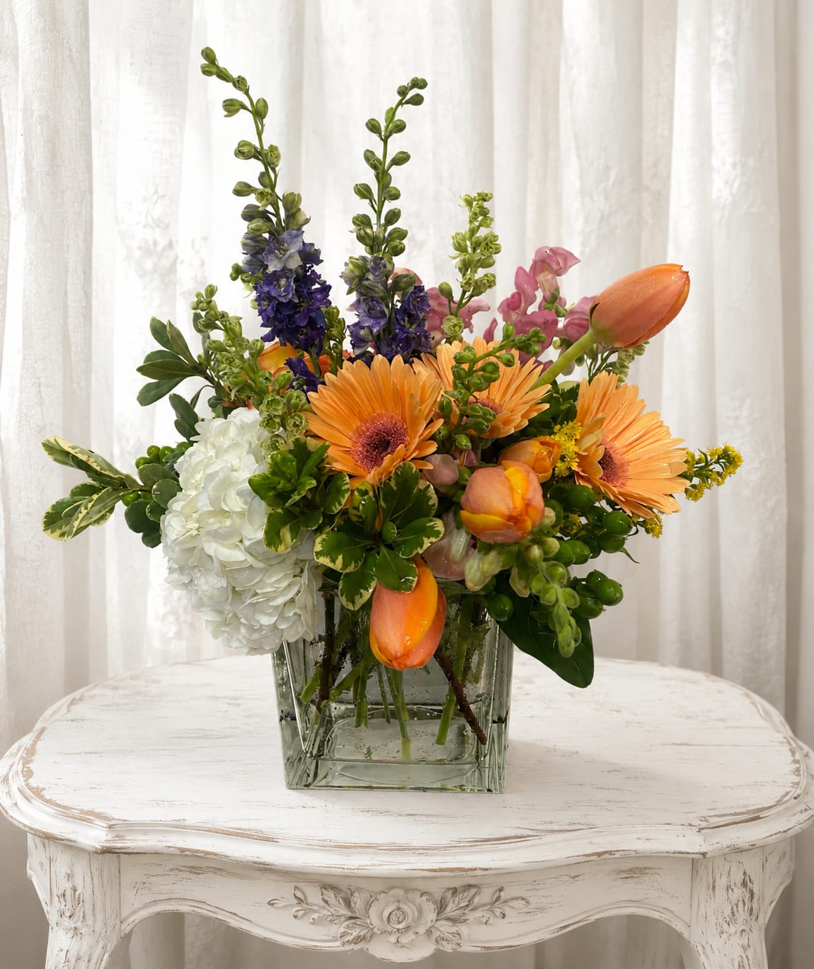 Tulips surprise - Bright, garden-fresh and joyfully unexpected, this spring bouquet is a Tulips surprise perfect for birthdays, thank-you gifts, or anyone who appreciates a lively burst of color. The arrangement places orange gerbera daisies and tulips front and center for immediate cheerful impact, while the white hydrangea cluster softens the palette and gives a lush, cloud-like contrast that feels celebratory and bright.  Tall purple and pink snapdragon spikes rise above the lower blooms to create a layered, garden-style silhouette that reads as both elegant and playful. Arranged in a clear square glass vase, the mix of textures-from the daisy centers to the rounded hydrangea heads and the tulip curves-makes the bouquet feel full and thoughtfully composed. It's the kind of gift that will make the person you're thinking of feel truly seen and celebrated; vase included and ready to display on a mantel, dining table, or bedside.   If any design elements are of major importance to your order, please include them in the florist instructions at checkout or contact us to ensure availability.