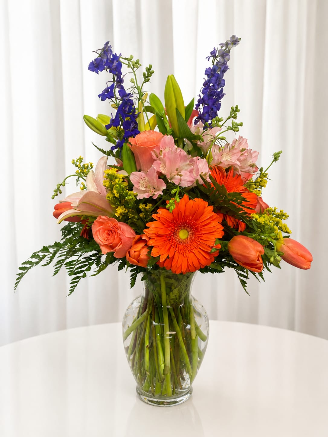 Bold and beautiful - Bright, lively, and unapologetically joyful - this mixed-color flower arrangement makes a bold statement for a birthday bouquet, congratulations gift, or any celebratory moment. Bright orange gerbera daisies and coral roses pop against cool blue delphinium-like spikes, while pink alstroemeria and tall lilies add layered height and a garden-style silhouette that will make the person you're thinking of feel truly celebrated.  Arranged upright in a clear glass vase, the composition is full and textured: round gerbera centers, soft rose petals, delicate alstroemeria blooms, and stately lily buds create contrast in shape and finish. The color palette of orange, pink and blue creates warm-versus-cool drama that reads joyful from across the room. Size and stem lengths are arranged to present an elegant vertical profile that works well on a dining table, office desk, or entry console.  Perfect for someone who loves vibrant color and a bit of showmanship, this arrangement sends a message of cheer and celebration without being over the top. Delivered ready to display in its vase, simple care - fresh water and a cool spot out of direct sun - will help the blooms stay bright for days.   If any design elements are of major importance to your order, please include them in the florist instructions at checkout or contact us to ensure availability.