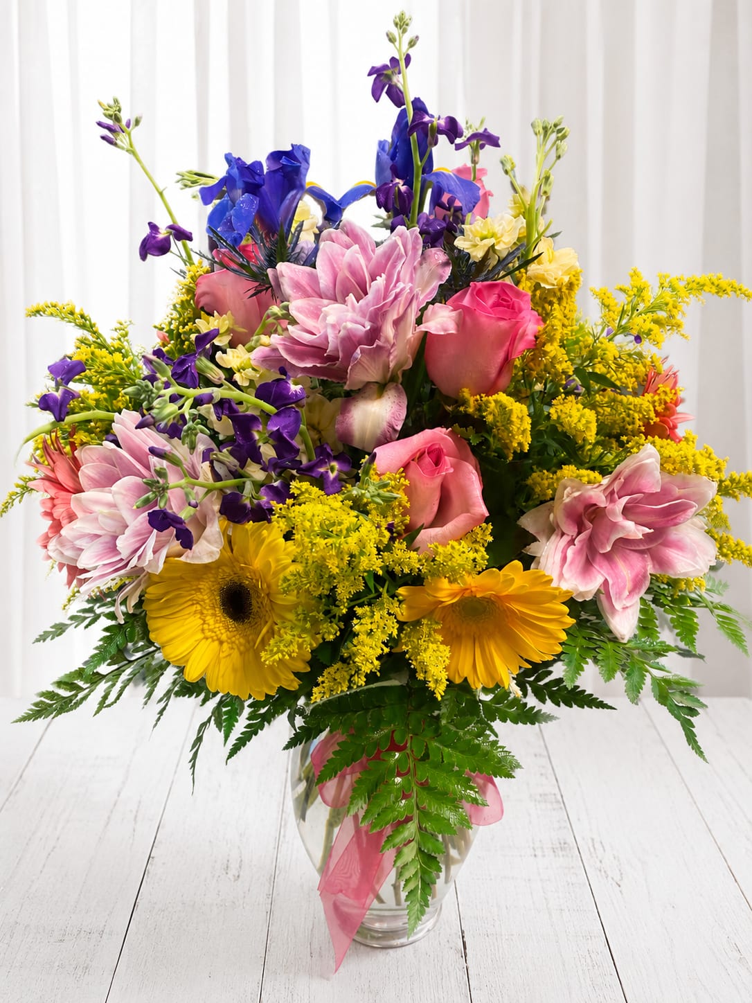 Be happy  - A lush, celebratory mixed bouquet that reads joyful at first glance - perfect as a birthday bouquet, congratulations arrangement, or a cheerful just-because gift. Pink roses and vivid purple accents rise above a rounded center of bright yellow daisy-like blooms, creating an energetic, uplifting palette that will make the person you're thinking of feel noticed and celebrated.  This large, full arrangement is densely layered for maximum presence and delivered in a clear glass vase tied with a soft pink ribbon. Alongside the roses and gerbera-style yellow blooms are classic white daisies and airy yellow filler that add texture and movement, so the bouquet looks abundant from every angle. Stems are arranged tall above the center to give height while keeping a generous, rounded silhouette.  Give this to someone who could use a bright moment - for birthdays, to mark an achievement, or simply to send sunshine on an ordinary day. Vase-ready for immediate display; refresh with fresh water every few days and trim stems at an angle to extend bloom life.   If any design elements are of major importance to your order, please include them in the florist instructions at checkout or contact us to ensure availability.