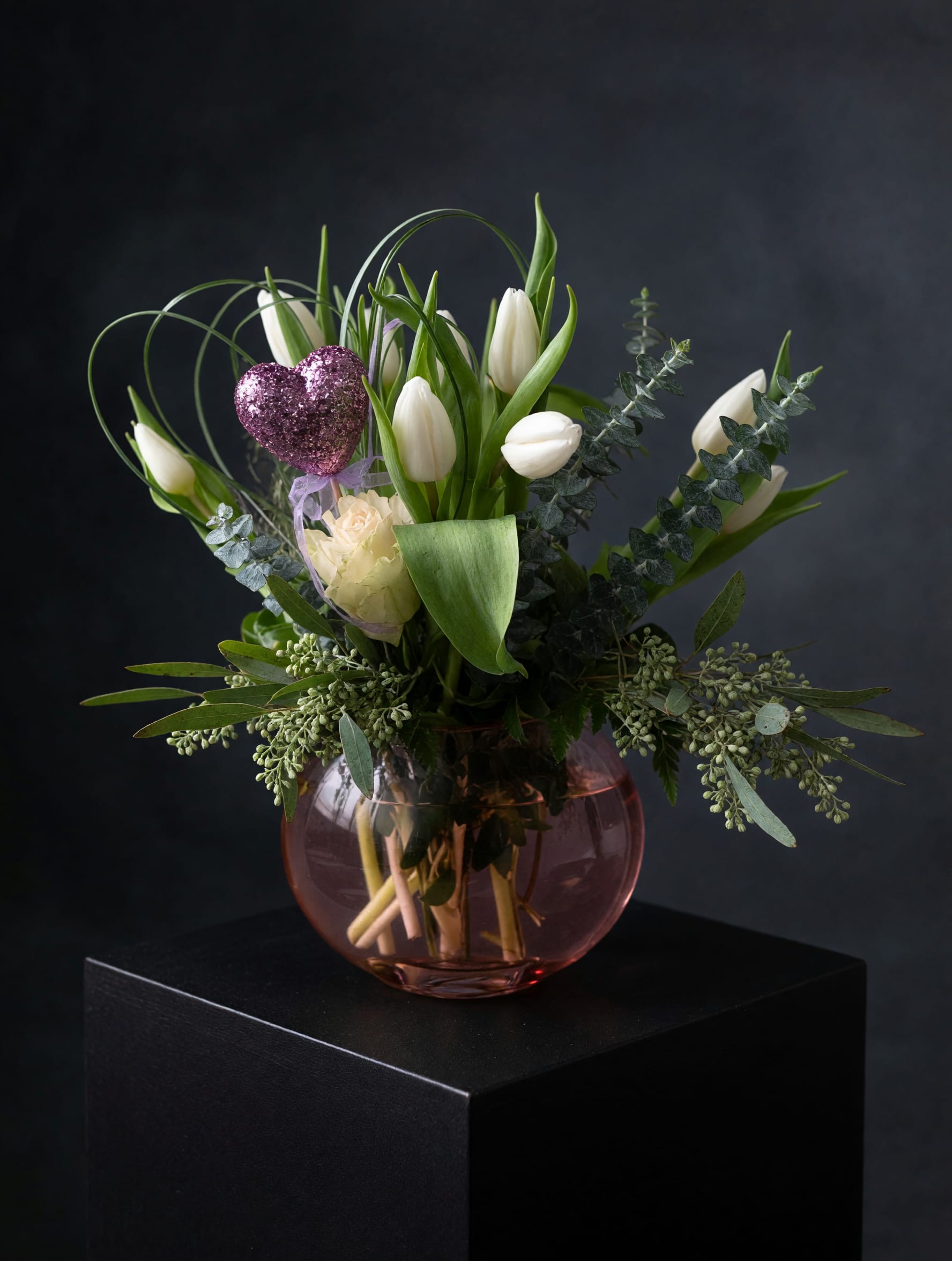White tulips arranged in a pink glass vase with a glittery heart pick