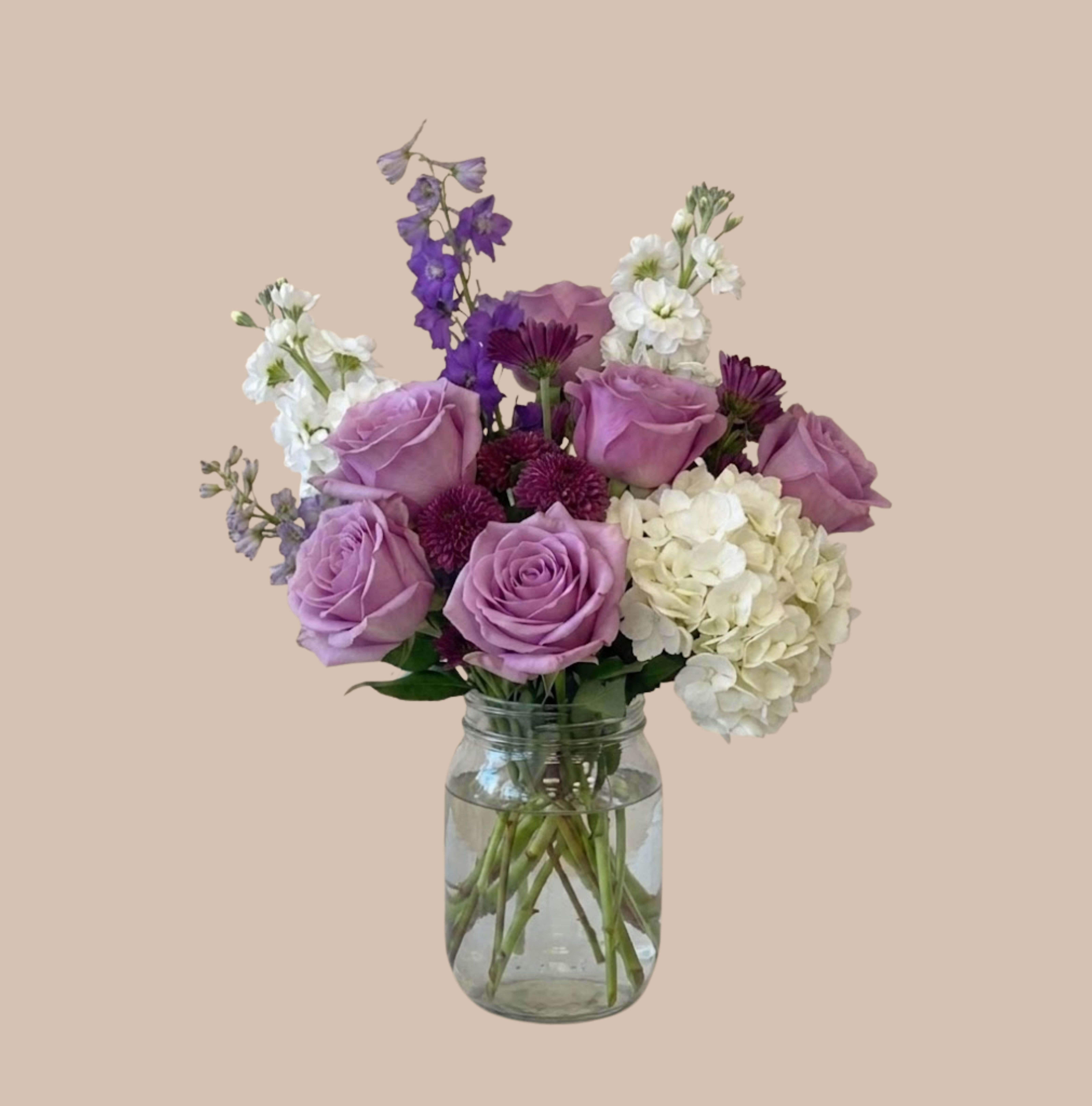 Stem &amp; Shadow Standard  - Lavender, white, and purple florals create a cool, romantic statement in this flower arrangement - a Stem &amp; Shadow Standard bouquet perfect for birthdays, anniversaries, thank-yous, or spring celebrations. The clear glass jar vase keeps the look fresh and casual while letting the pale and deep purples take center stage, so the person you're surprising will feel noticed and genuinely celebrated.  Rounded rose heads mingle with airy vertical delphinium spikes and small dark plum accent blooms for a garden-style silhouette that's at once soft and lively. Colors are a blend of purple, white, and pink tones with bright white hydrangea lifting the richer hues; the mix of textures adds visual movement and keeps the arrangement feeling natural rather than formal.  This everyday bouquet arrives arranged in a clear glass jar - an easy, display-ready gift that fits on a mantel, bedside table, or kitchen counter. No special care is required beyond fresh water and a cool spot away from direct sun; choose this arrangement when you want a thoughtful, elegant surprise that reads as both intimate and joyful.