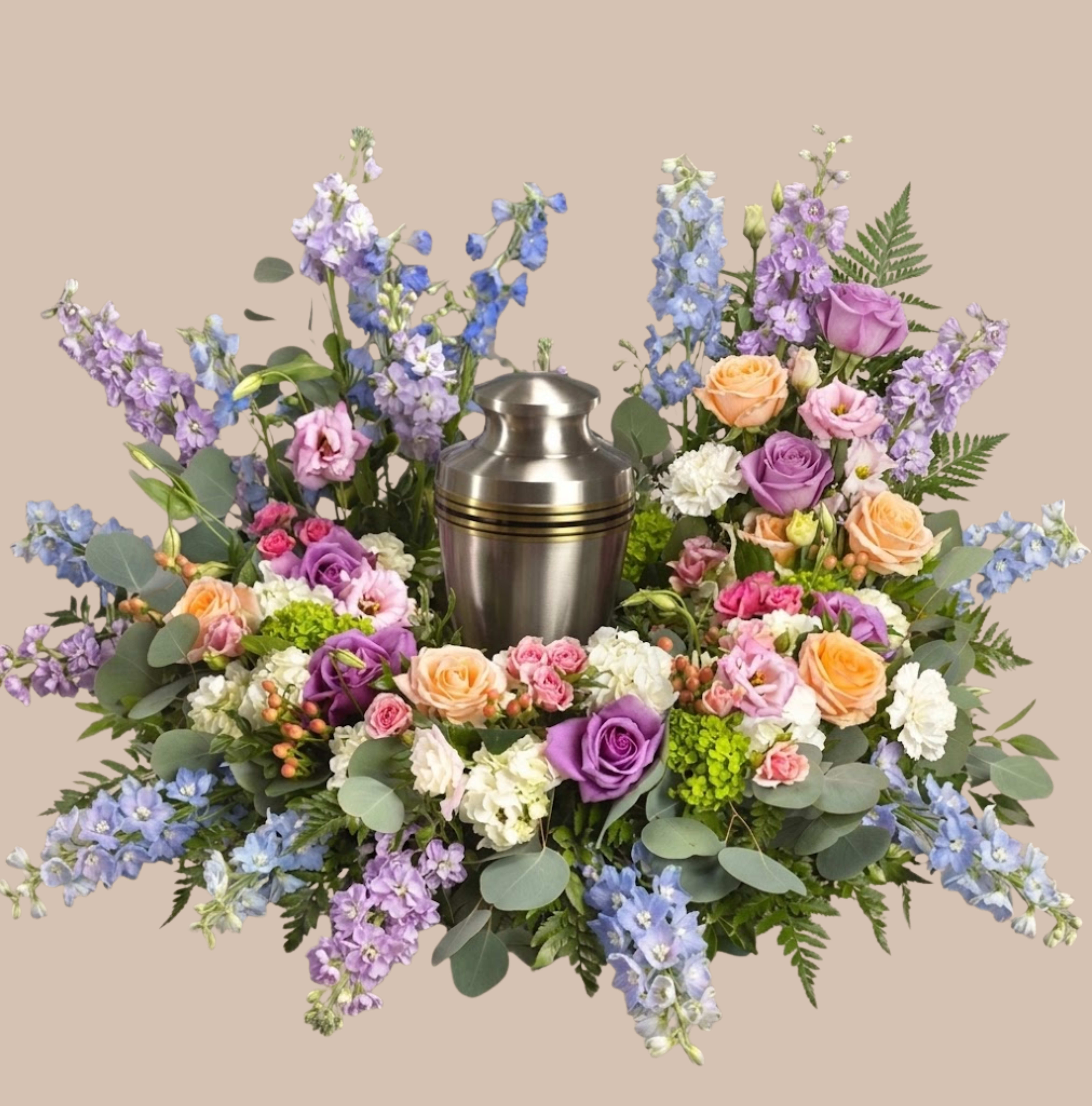 Pastel Clouds Tribute - A cloud-like collar of pastel blooms surrounds a central urn to create a serene sympathy arrangement perfect for funerals and memorial services. This urn surround-designed to sit closely around a standard black or metal urn-reads as a formal, comforting tribute that gently honors the person you're remembering, making them feel respected and deeply remembered.  Soft lavenders, pale pinks and peaches mingle with whites, touches of yellow and deeper blue accents for depth. Large blue hydrangea clusters anchor the design while white roses, white calla lilies, spray roses, carnations, and tall delphinium add layered texture and a graceful vertical line. Eucalyptus, fern and mixed greenery form a broad, low base so the arrangement is full and symmetrical, visible and balanced from every angle.  Made specifically as an urn surround for a memorial setting, this piece is sized and arranged to photograph well at a service and present a peaceful focal point at a cemetery chapel or funeral home. Lightweight care guidance: keep flowers in a cool, shaded area until the service; the design is crafted to maintain its shape and dignity throughout viewing and ceremony.