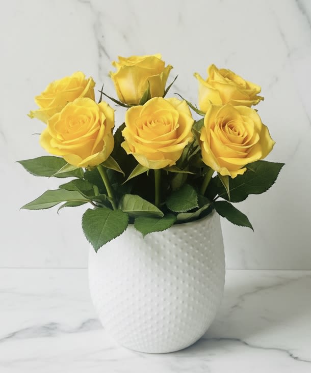 golden roses - Six bright yellow roses gathered into a compact, sunlit cluster make an immediate cheerful statement-perfect as a birthday bouquet, a thank-you arrangement, or a just-because surprise to lift someone's day. The warm yellow petals read joyful and optimistic, while the neat, modern presentation keeps the look fresh and approachable for everyday gifting or spring celebrations.  Displayed in a white textured vase, the open, full blooms sit low and close for an elegant, sculptural silhouette that works on tabletops, entry consoles, or office desks. Accents of green foliage and touches of white quietly balance the color story without stealing focus. Care is simple: keep the vase filled with fresh water, trim stems every few days, and enjoy six long-lasting roses arranged for immediate impact.