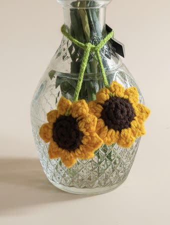 Hanging crocheted Sunflowers - Handmade crocheted sunflowers bring a sunny, rustic charm to any room-perfect as a cheerful birthday bouquet alternative or a thoughtful "just because" gift for someone who loves handmade decor. This hanging crocheted sunflower arrangement pairs two bright yellow and orange sunflower blooms with deep black-brown centers, arranged to sit against the clear patterned glass vase so the flowers read like warm, perpetual blooms. The casual, craft-like look makes it ideal for kitchen windows, a cozy reading nook, or a dorm room where low-maintenance charm matters.  The clear glass vase shows off the clean silhouette of the stems and keeps the focus on the textured yarn flowers; a simple green yarn tie around the neck adds a finished, rustic touch. Because these are crocheted, they offer long-lasting color and texture without watering or special care-she'll enjoy the look of fresh sunflowers every day. Choose this for housewarmings, teacher gifts, or a bright accent to celebrate spring or summer moods year-round.
