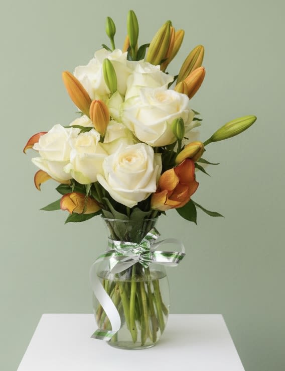 Lilies and Roses - Creamy white roses meet warm orange and sunny yellow lilies in a compact, upright bouquet that feels both fresh and refined-perfect as a birthday bouquet, thank-you arrangement, or a bright sympathy or thinking-of-you gift. The mix of open roses and unopened lily buds gives the design a layered, hopeful quality that reads as gentle romance for an anniversary, cheerful support for a new baby or graduation, and a polished choice for Father's Day or retirement celebrations.  Arranged in a clear glass vase and finished with a white-and-green satin ribbon, the display is neat, rounded, and easy to place on a mantel, desk, or bedside table. Expect creamy white petals, orange lily blooms and buds, and touches of green foliage assembled for a balanced silhouette; sizing and vase dimensions follow our standard bouquet proportions for table-friendly presence. Simple care-fresh water and a cool spot-keeps blooms lasting longer, making this an elegant every-season gift for weddings, Passover, Rosh Hashanah, Mother's Day, or just because.
