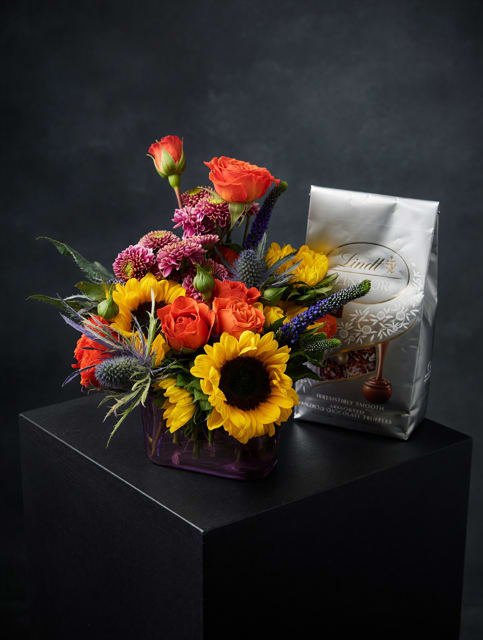 Sunflowers Mix with Lindor Chocolates - Bright, cheerful sunflowers steal the show in this mixed bouquet designed for birthdays, "just because" surprises, or a thoughtful Father's Day treat. The arrangement reads like a celebration-sunflowers paired with orange roses and pink‑purple chrysanthemums create a warm, joyful palette while spiky blue‑purple accents add lift and texture that catch the eye. It's the kind of gift that makes the person you're thinking of feel seen and celebrated.  Arranged in a purple glass vase, the compact bouquet is polished and easy to display on a kitchen counter or bedside table. The mix includes sunflowers, roses, chrysanthemums, and other seasonal blooms in yellow, orange and mixed purple tones; a silver Lindt chocolate bag is presented alongside in a matching gift bag for an extra sweet touch. Perfect for get well wishes, thank yous, new baby visits, anniversaries or simple thinking-of‑you moments-this set arrives ready to give and brings bright color and comforting scent into any room.