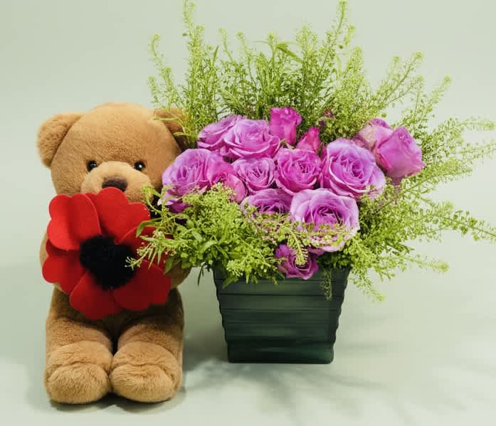 Teddy bear beside a vase of purple roses and greenery