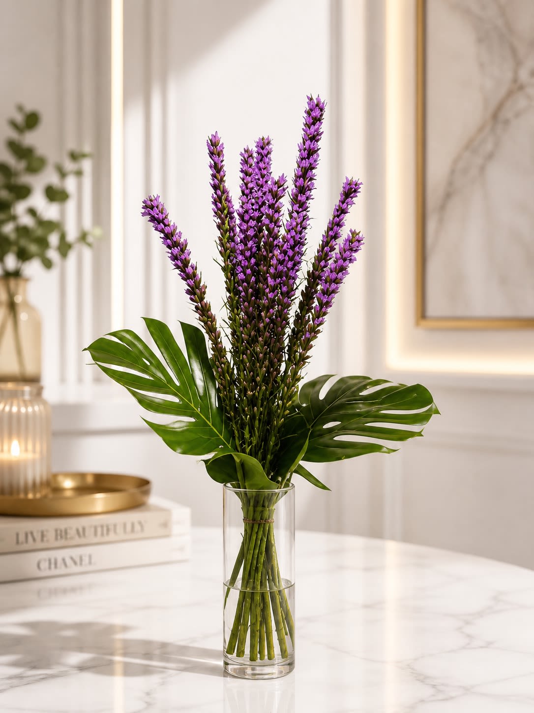 Violet Sol - A sleek and modern floral design featuring vibrant purple liatris accented with lush tropical monstera leaves in a tall clear glass cylinder vase. Violet Sol is designed with clean lines and dramatic height, creating a striking contemporary arrangement that feels elegant, artistic, and effortlessly upscale. The rich purple blooms bring bold color and texture while the tropical greenery adds movement and a fresh modern touch, making this arrangement perfect for birthdays, office spaces, thank-you gifts, sympathy gestures, or sophisticated home décor.  To provide the freshest arrangement possible, certain flowers or greenery may occasionally be substituted with blooms or foliage of equal or greater value while preserving the overall modern tropical aesthetic and purple color palette of the design. Similar premium seasonal florals and contemporary greenery may be used based on availability. Vase styles may also vary slightly depending on inventory, but the arrangement will always be presented in a comparable modern premium container that complements the overall look and value.