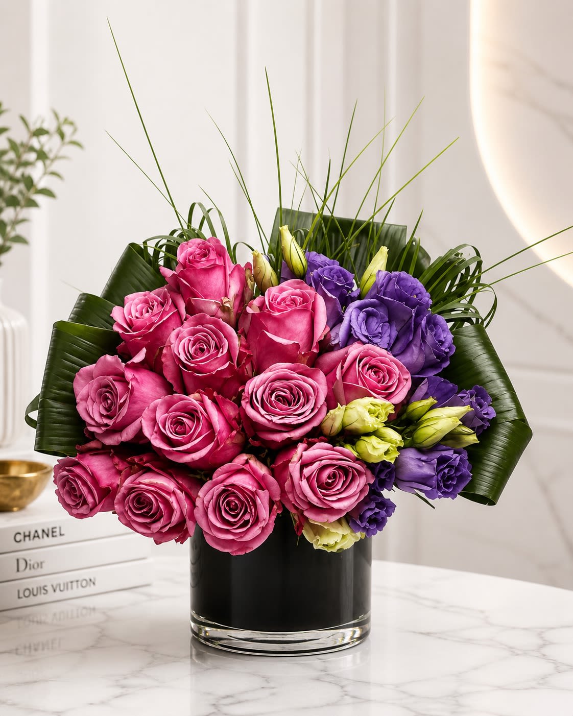 Velvet Amore - A luxurious modern arrangement featuring premium hot pink roses accented with rich purple lisianthus, sculpted tropical foliage, and contemporary greenery in a sleek black glass cylinder vase. Velvet Amore is designed with bold romantic tones and elegant texture, creating a striking upscale statement that feels both glamorous and sophisticated. The vibrant pink roses bring depth and passion while the soft purple lisianthus adds contrast and dimension, making this arrangement perfect for birthdays, anniversaries, romantic gestures, celebrations, or luxury everyday décor.  To ensure the freshest design possible, certain flowers or greenery may occasionally be substituted with blooms of equal or greater value while maintaining the overall color palette, premium style, and modern aesthetic of the arrangement. Similar luxury roses, lisianthus varieties, or tropical foliage may be used based on seasonal availability. The container may also vary slightly depending on inventory, but the arrangement will always be created in a comparable modern premium vase that complements the overall design and presentation.