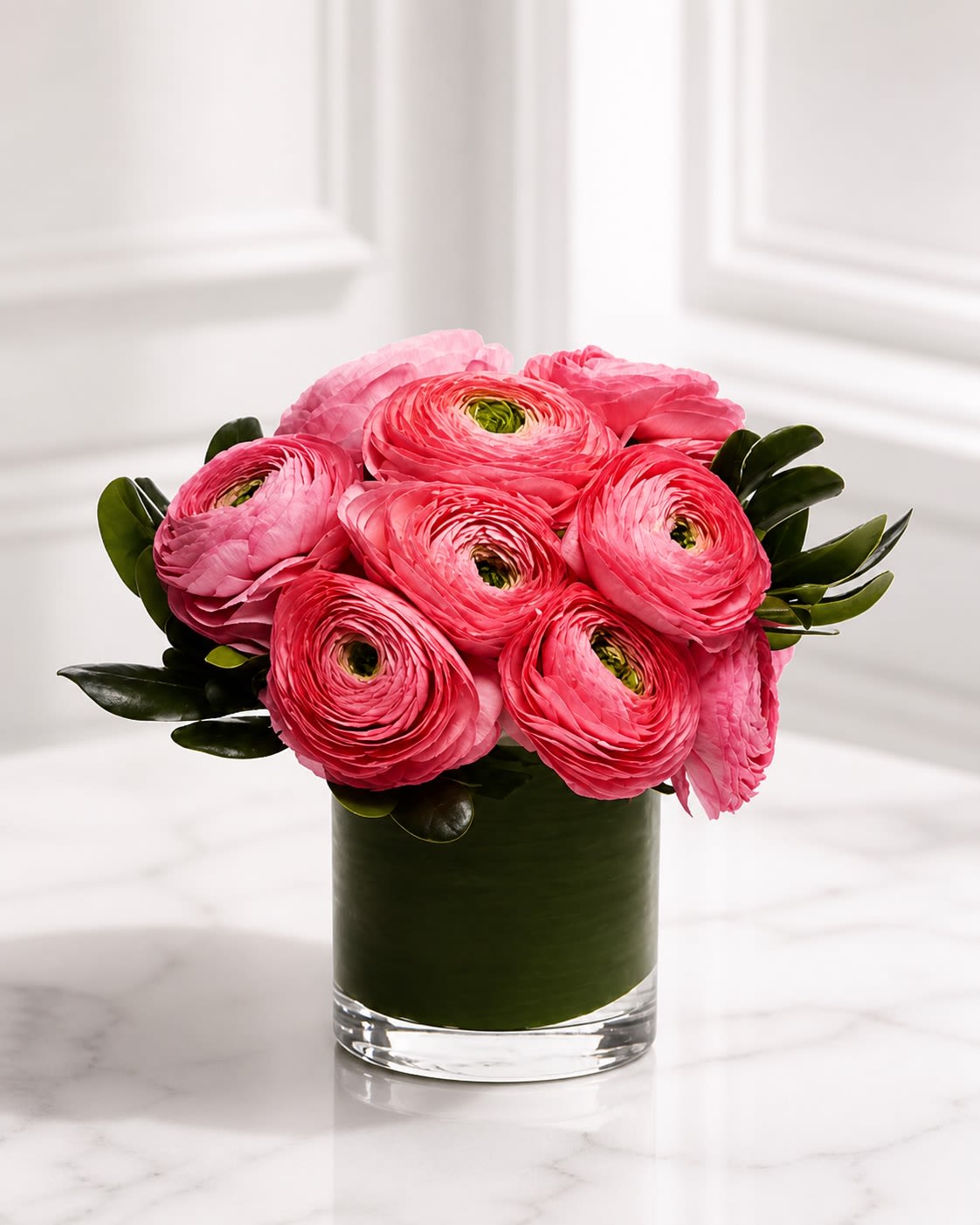 Rosé Ranunculus - A chic luxury arrangement featuring premium hot pink and soft blush ranunculus designed in a modern leaf-lined glass cylinder vase with rich glossy greenery accents. Rosé Ranunculus highlights the extraordinary layered petals and delicate spiral texture that make ranunculus one of the most sought-after luxury blooms in floral design. The vibrant pink tones paired with the sleek contemporary presentation create a romantic yet fashion-forward arrangement perfect for birthdays, celebrations, thank-you gifts, anniversaries, or elegant everyday décor.  Designed with premium seasonal ranunculus and modern foliage, this arrangement offers a lush garden-inspired appearance with a clean upscale aesthetic. Due to seasonal availability, certain flowers or greenery may occasionally be substituted with blooms of equal or greater value while preserving the overall color palette, luxurious texture, and modern style of the arrangement. The leaf-lined cylinder vase may also vary slightly depending on inventory, but the design will always maintain its elevated contemporary presentation.