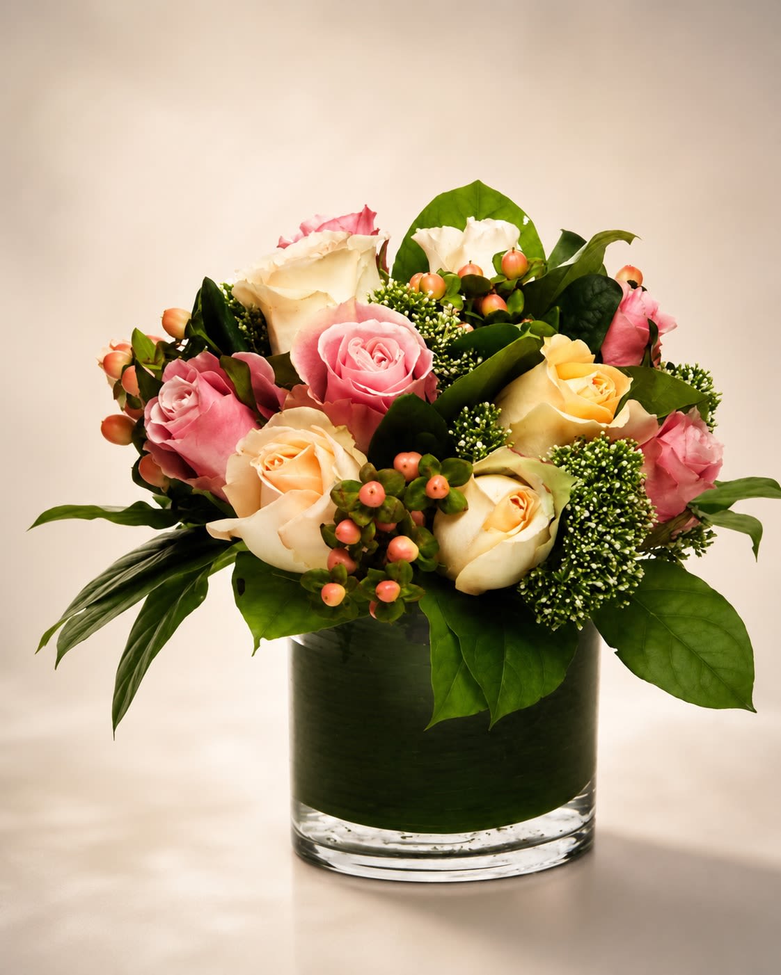 Golden Rosé Garden - A refined luxury arrangement featuring premium pink, ivory, and soft champagne roses accented with hypericum berries, textured seasonal greenery, and contemporary foliage in a modern leaf-lined glass cylinder vase. Golden Rosé Garden blends romantic garden-inspired tones with a sleek modern presentation, creating an arrangement that feels elegant, warm, and effortlessly upscale. The soft pastel palette paired with rich green textures gives this design a fresh couture floral aesthetic perfect for birthdays, anniversaries, thank-you gifts, celebrations, or sophisticated everyday décor.  Designed using premium seasonal roses and luxury greenery, this arrangement offers a lush balanced composition with soft romantic color tones and modern styling. Certain flowers, berries, or foliage may occasionally be substituted with blooms of equal or greater value depending on seasonal availability while preserving the overall color palette, luxurious feel, and elevated presentation of the arrangement. The leaf-lined cylinder vase may also vary slightly based on inventory, but the overall modern aesthetic and premium quality will always be maintained.