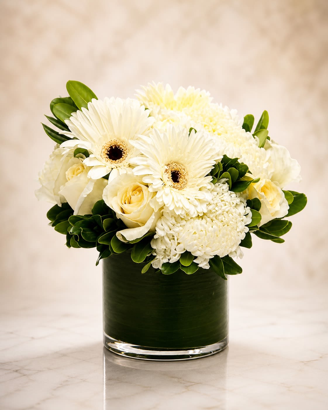 White Velvet Garden - A refined all-white floral arrangement designed with elegant white gerbera daisies, premium roses, cushion mums, and seasonal textured blooms gathered in a modern clear cylinder vase wrapped with rich green foliage lining. White Velvet Garden brings together softness, texture, and clean luxury styling in a timeless monochromatic palette that feels both calming and sophisticated.  This arrangement is designed in a lush rounded style with layered white florals and polished greenery for an upscale presentation suitable for sympathy occasions, birthdays, elegant home décor, thank-you gifts, or luxury everyday floral styling. The combination of crisp white blooms and deep green accents creates a fresh and serene design with a modern floral aesthetic.  Due to seasonal and market availability, certain flowers may occasionally be substituted with blooms of equal or greater value while maintaining the overall color palette, fullness, and luxurious garden-inspired style of the arrangement. Vase style and foliage lining may also vary slightly depending on availability while preserving the same upscale presentation.