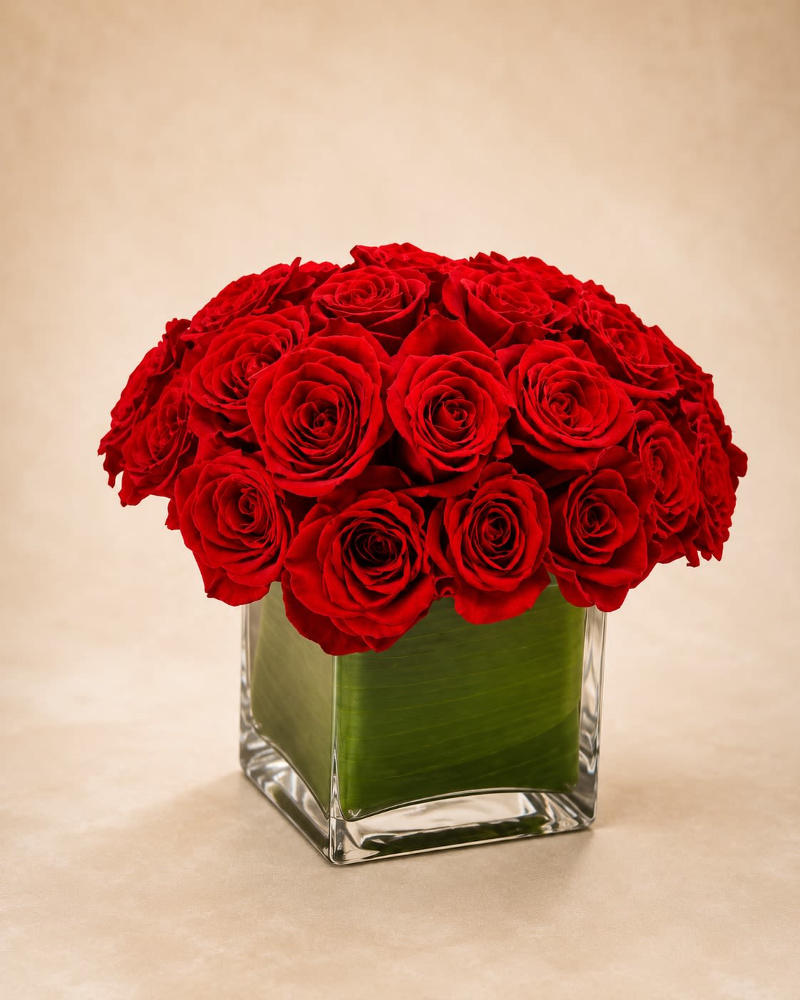 Crimson Couture Roses - A luxurious statement arrangement featuring premium long-stem red roses artfully designed in a modern clear glass cube vase lined with rich green foliage for a polished contemporary finish. Crimson Couture Roses is designed in a lush dome silhouette that showcases the timeless beauty and romance of classic red roses with an elevated luxury presentation.  This arrangement is crafted with tightly clustered premium roses to create a full, velvety appearance perfect for romantic occasions, anniversaries, Valentine’s Day, milestone celebrations, or elegant luxury gifting. The clean cube vase paired with layered tropical leaf lining gives the design a refined modern aesthetic while allowing the vibrant crimson blooms to remain the focal point.  Due to seasonal and market availability, rose varieties or shades may occasionally vary slightly while maintaining the same rich red color palette and luxurious style. Vase shape and foliage lining may also vary based on availability while preserving the arrangement’s upscale presentation and fullness.