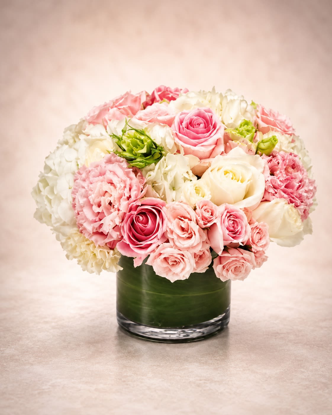 Rosé Garden Luxe - A soft and romantic luxury floral arrangement featuring premium pink and ivory roses, blush spray roses, white hydrangea, delicate carnations, and seasonal accent blooms artfully arranged in a modern clear cylinder vase lined with rich green foliage. Rosé Garden Luxe is designed with a lush rounded silhouette that blends elegant textures and soft feminine tones into an upscale garden-inspired presentation.  This arrangement brings together layered premium blooms in shades of soft pink, ivory, and creamy blush to create a timeless floral design perfect for birthdays, baby showers, anniversaries, sympathy gestures, thank-you gifts, or elegant everyday décor. The combination of full hydrangea, garden-style roses, and delicate textural blooms creates a rich luxurious look while maintaining a soft romantic feel.  Due to seasonal and market availability, certain flowers may occasionally be substituted with blooms of equal or greater value while preserving the arrangement’s color palette, fullness, and overall luxury garden aesthetic. Vase style and foliage lining may also vary slightly depending on availability while maintaining the same elegant presentation.