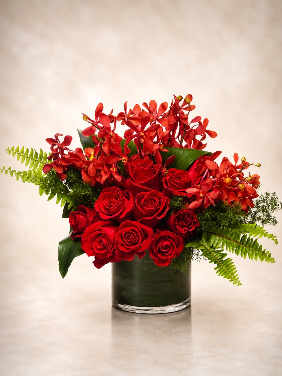 Scarlet Orchid Affair - A bold luxury floral arrangement featuring premium red roses paired with vibrant red mokara orchids, lush tropical foliage, and fresh textured greenery arranged in a modern leaf-lined cylinder vase. Scarlet Orchid Affair is designed with a rich contemporary tropical style that combines classic romance with exotic elegance for a dramatic upscale presentation.  This arrangement showcases layers of velvety red roses surrounded by striking orchid blooms and flowing greenery to create a luxurious statement piece perfect for anniversaries, romantic celebrations, grand gestures, upscale events, or sophisticated gifting. The contrast of vivid crimson tones against tropical textures gives the design a lavish couture-inspired aesthetic with a modern exotic touch.  Due to seasonal and market availability, certain orchid varieties, greenery, or rose shades may occasionally vary while maintaining the same rich red palette, tropical elegance, and overall fullness of the arrangement. Vase style and foliage lining may also vary slightly depending on availability while preserving the arrangement’s luxurious presentation.