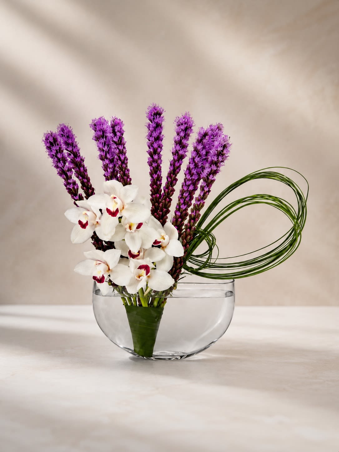 Orchid Amethyst Zen - A striking contemporary floral design featuring elegant white cymbidium orchids paired with vibrant purple liatris blooms, artfully accented with modern looping grass in a sleek clear glass bowl vase. Orchid Amethyst Zen blends modern minimalism with exotic luxury, creating a sophisticated statement piece full of texture, movement, and artistic elegance.  The rich violet tones of the liatris beautifully contrast against the crisp white orchids, while the sculptural grass accents add a refined contemporary touch perfect for upscale interiors and luxury gifting. This arrangement is ideal for birthdays, corporate gifting, modern home décor, thank-you gestures, wellness spaces, or anyone who appreciates artistic floral design.  Due to seasonal and market availability, certain orchid blooms, accent florals, or greenery may occasionally vary while preserving the arrangement’s elegant color palette, contemporary styling, and luxurious presentation. Vase style may also vary slightly depending on availability while maintaining the same modern aesthetic.
