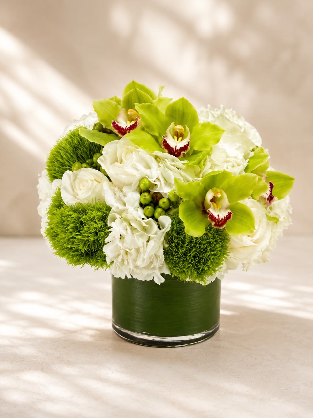 Emerald Orchid Luxe - A breathtaking luxury arrangement featuring vibrant green cymbidium orchids, lush white hydrangeas, premium white roses, textured green dianthus blooms, and fresh hypericum berries artfully arranged in a modern leaf-lined glass vase. Emerald Orchid Luxe blends crisp ivory tones with vivid botanical greens for a fresh, contemporary floral design that feels both sophisticated and fashion-forward.  This elegant arrangement symbolizes harmony, prosperity, renewal, and refined beauty, making it a perfect choice for upscale gifting, birthdays, congratulations, corporate events, sympathy occasions, or luxurious home décor. The striking green orchids create a bold focal point while the soft hydrangeas and roses add fullness and timeless elegance.  Due to seasonal and market availability, specific orchid shades, berry accents, or specialty blooms may vary slightly while maintaining the arrangement’s luxurious green-and-white palette and modern aesthetic. Vase style may occasionally vary depending on availability while preserving the same upscale presentation.