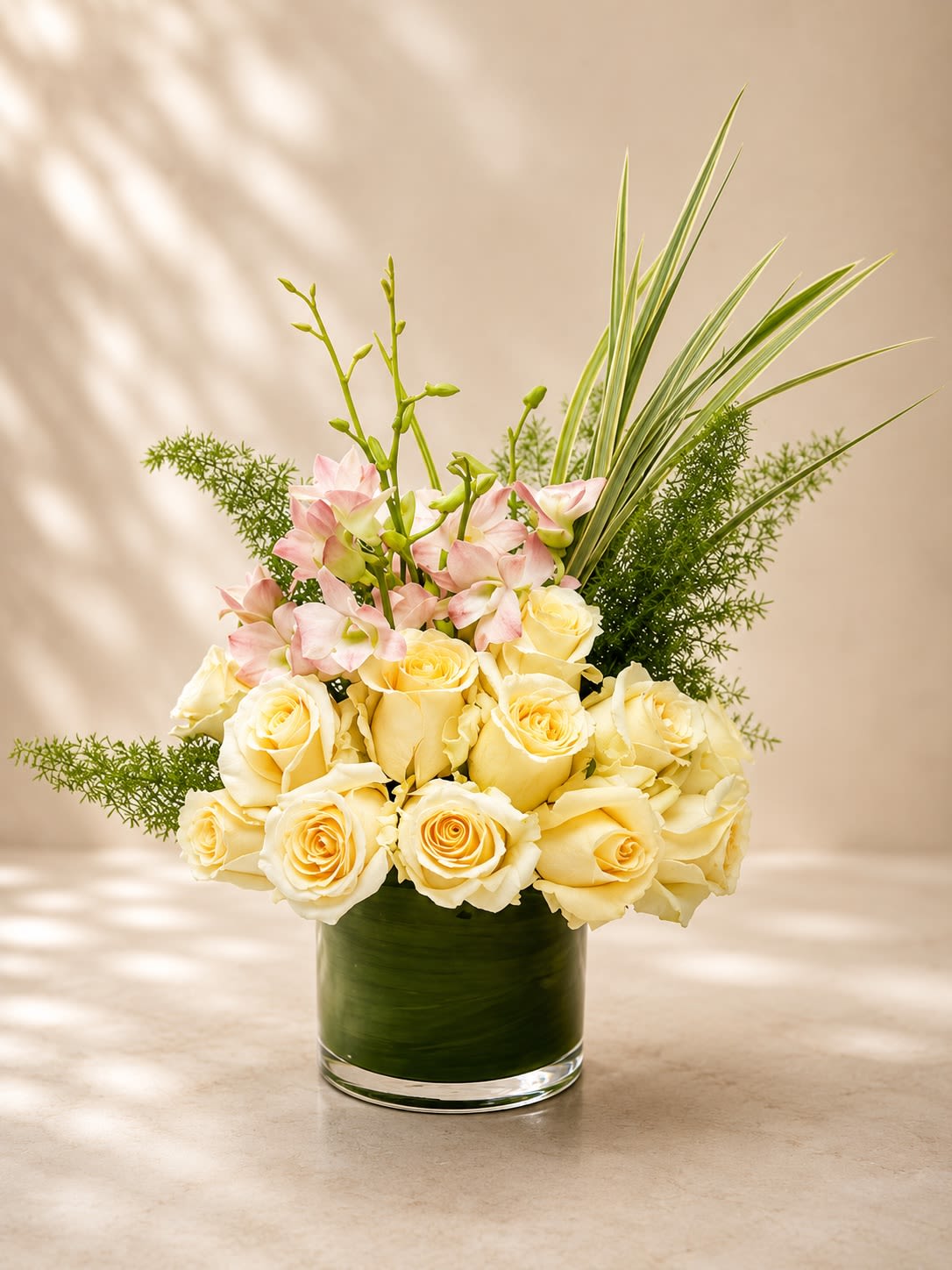 Ivory Garden Elegance - A refined and luxurious floral arrangement featuring creamy ivory roses, delicate blush orchids, lush textured greenery, and modern tropical foliage designed in a sleek leaf-lined glass vase. Ivory Garden Elegance captures timeless sophistication with a fresh botanical feel, blending soft romantic blooms with contemporary design elements for an effortlessly upscale presentation.  This arrangement symbolizes grace, purity, admiration, and serenity, making it a beautiful choice for sympathy gestures, anniversaries, birthdays, thank-you gifts, corporate gifting, or elegant home décor. The soft ivory tones paired with airy greenery and delicate orchid accents create a design that feels both tranquil and couture-inspired.  Due to seasonal and market availability, orchid varieties, greenery textures, and rose shades may vary slightly while preserving the arrangement’s luxurious ivory and blush palette and overall elegant style. Vase style may occasionally vary depending on availability while maintaining the same premium presentation.