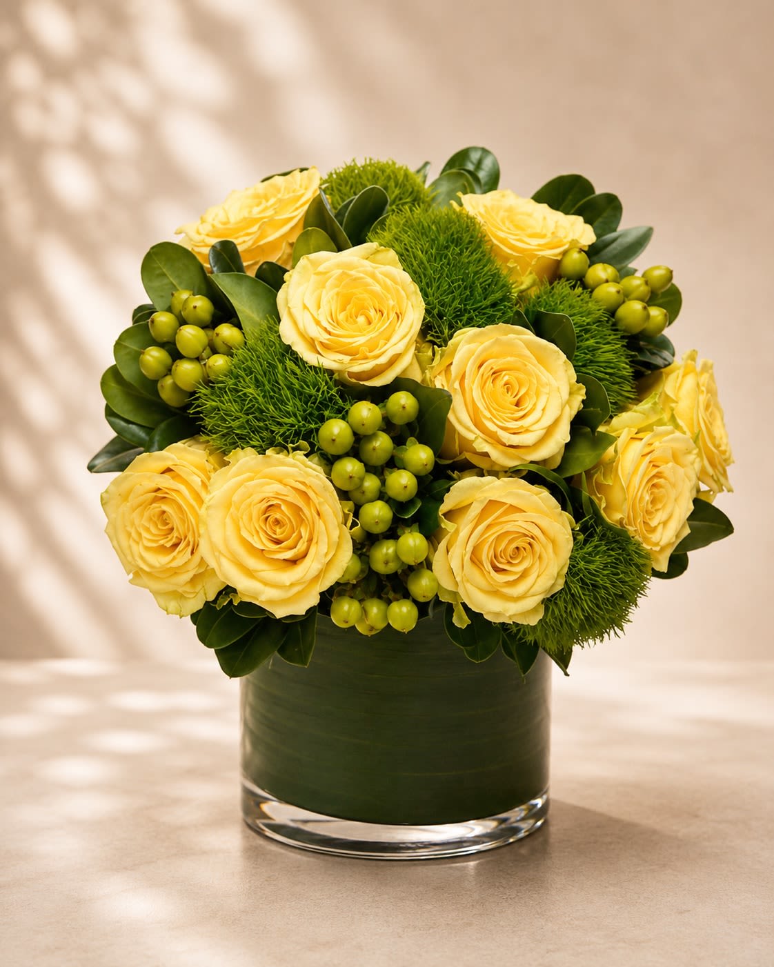 Citrus Grove Couture - A vibrant and refreshing floral arrangement featuring soft lemon-yellow roses accented with lush green dianthus, hypericum berries, and rich glossy foliage arranged in a sleek leaf-lined glass vase. Citrus Grove Couture combines modern elegance with lively botanical textures, creating a design that feels fresh, luxurious, and naturally uplifting.  Yellow roses symbolize joy, friendship, optimism, and happiness, while the vibrant green accents add a contemporary garden-inspired touch full of energy and sophistication. This arrangement is perfect for birthdays, congratulations, thank-you gifts, housewarmings, corporate gifting, or simply bringing brightness and positivity into any space.  Due to seasonal and market availability, greenery textures and berry accents may vary slightly while preserving the arrangement’s signature yellow-and-green palette and luxurious modern style. Vase style may occasionally vary depending on availability while maintaining the same premium presentation.