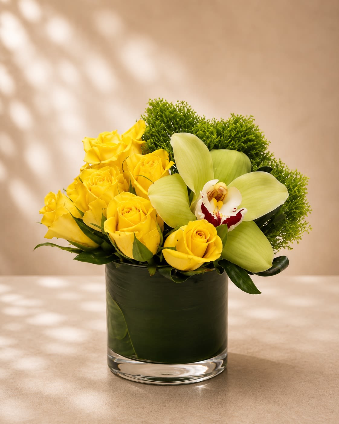 Lemon Orchard Elegance - A bright and refreshing floral arrangement featuring vibrant yellow roses, an elegant green cymbidium orchid, lush textured greenery, and modern tropical foliage arranged in a sleek leaf-lined glass vase. Lemon Orchard Elegance combines cheerful color with contemporary sophistication, creating a design that feels fresh, uplifting, and luxuriously refined.  Yellow roses symbolize joy, friendship, optimism, and happiness, while the graceful cymbidium orchid adds an elevated botanical touch representing beauty, strength, and elegance. The rich green textures and clean modern styling make this arrangement perfect for birthdays, congratulations, thank-you gifts, get well wishes, corporate gifting, or stylish everyday décor.  Due to seasonal and market availability, orchid varieties and greenery textures may vary slightly while preserving the arrangement’s signature yellow-and-green palette and luxurious modern garden style. Vase style may occasionally vary depending on availability while maintaining the same premium presentation.