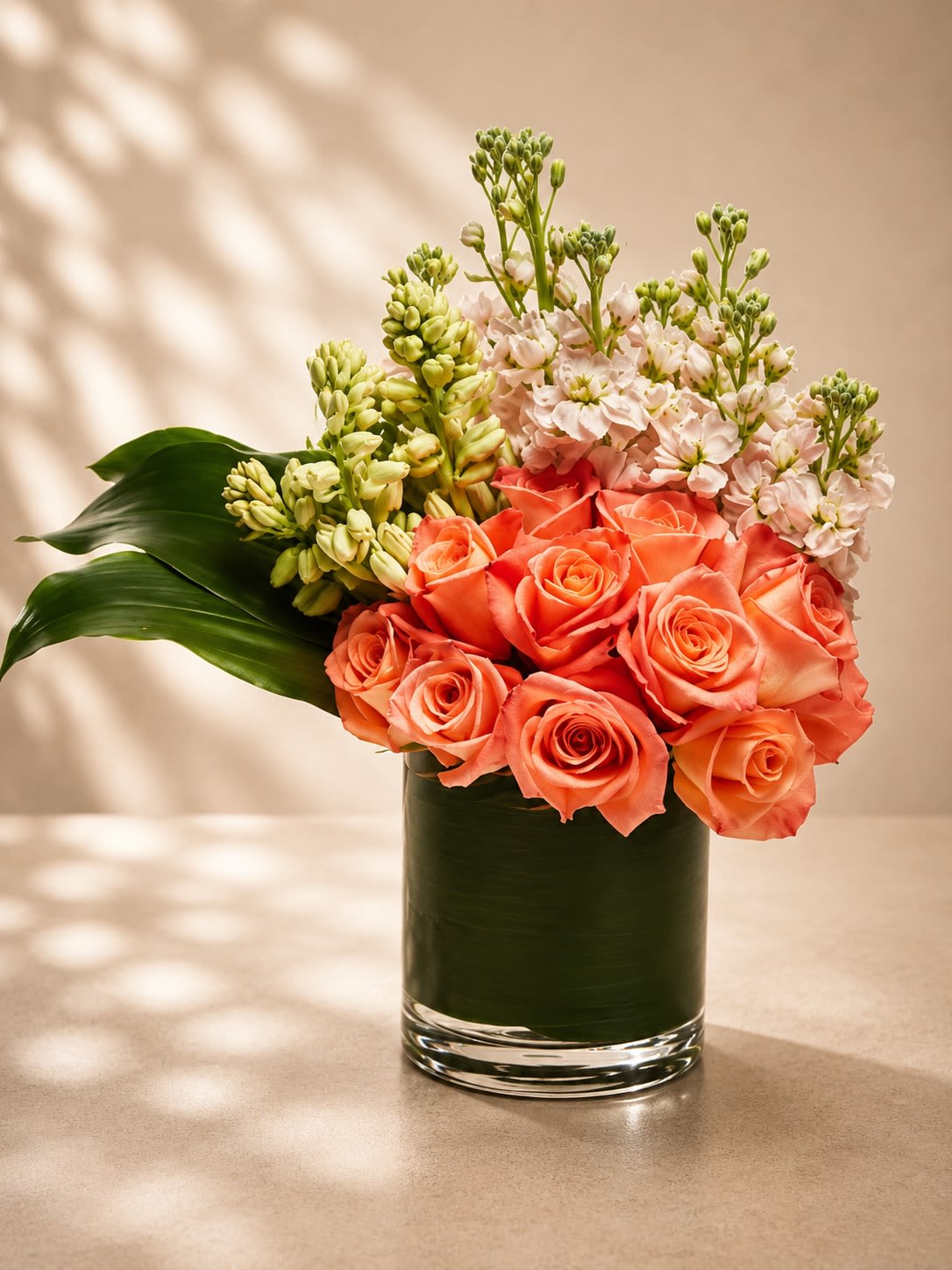 Coral Blossom Garden - A soft and cheerful floral arrangement featuring radiant coral roses accented with fragrant stock flowers, fresh green hyacinth blooms, and lush tropical foliage arranged in a modern leaf-lined glass vase. Coral Blossom Garden blends romantic warmth with fresh garden textures, creating a design that feels joyful, elegant, and naturally sophisticated.  Coral roses symbolize happiness, admiration, enthusiasm, and affection, while the delicate stock flowers add softness and a light floral fragrance that enhances the arrangement’s fresh garden-inspired feel. The vibrant peach and coral tones paired with crisp greenery make this arrangement perfect for birthdays, celebrations, thank-you gifts, anniversaries, Mother’s Day, or brightening any room with uplifting elegance.  Due to seasonal and market availability, stock flower shades and greenery textures may vary slightly while preserving the arrangement’s signature coral-and-blush palette and luxurious garden style. Vase style may occasionally vary depending on availability while maintaining the same premium presentation.