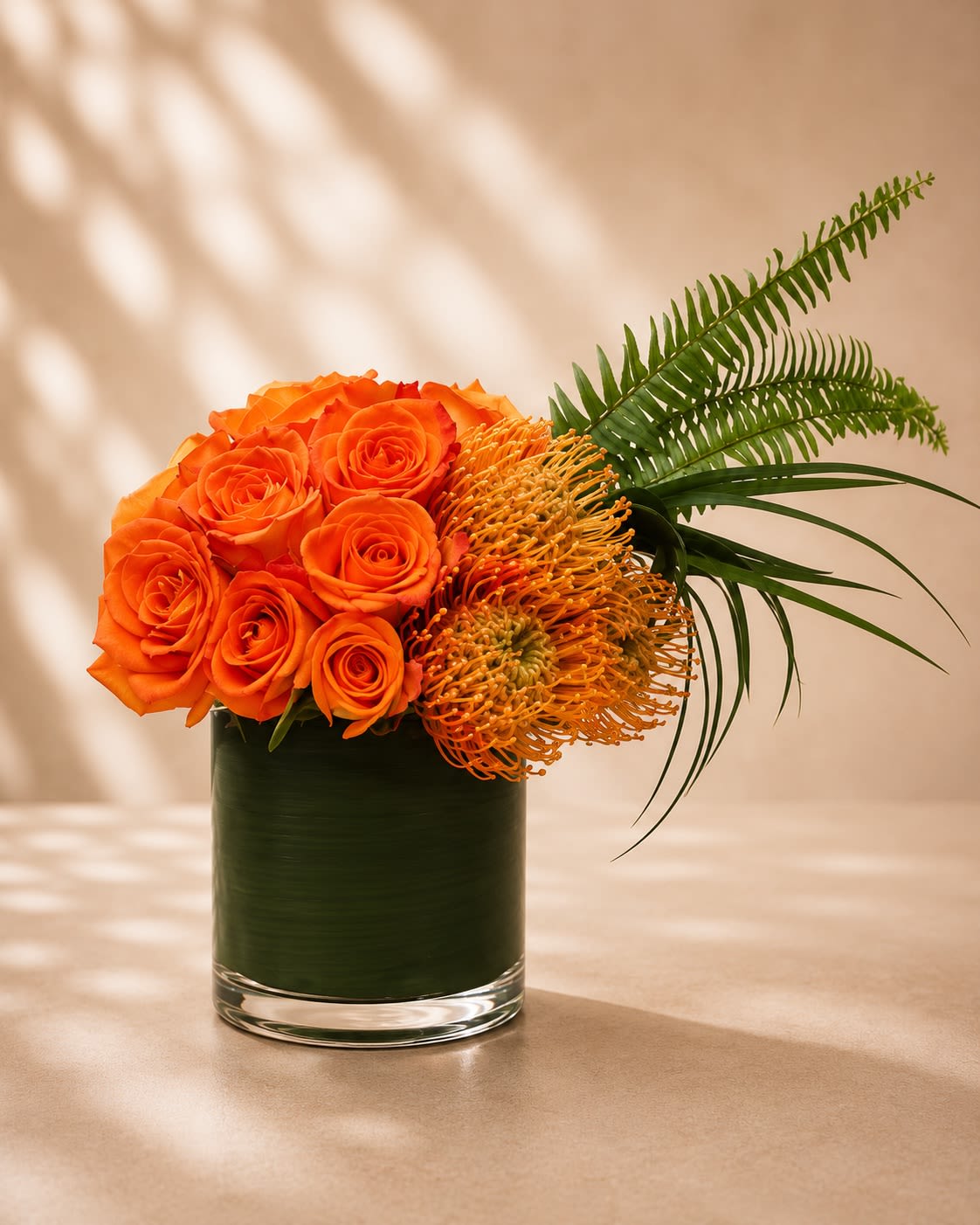 Sunset Ember Luxe - A bold and modern floral arrangement featuring vibrant orange roses paired with striking orange pincushion protea and sculptural tropical greenery in a sleek leaf-lined glass vase. Sunset Ember Luxe captures the warmth of a glowing sunset with rich citrus tones and dramatic textures, creating a design that feels both contemporary and luxurious.  Orange roses symbolize passion, excitement, admiration, and energy, while the exotic pincushion protea adds a unique artistic texture that makes this arrangement stand out beautifully. Accented with flowing tropical foliage and elegant fern details, this design is perfect for celebrations, birthdays, congratulations, thank-you gifts, housewarmings, or anyone who loves bold modern florals.  Due to seasonal and market availability, protea varieties and greenery may vary slightly while maintaining the arrangement’s signature warm sunset-inspired palette and upscale tropical style. Vase style may occasionally vary depending on availability while preserving the same elegant presentation.