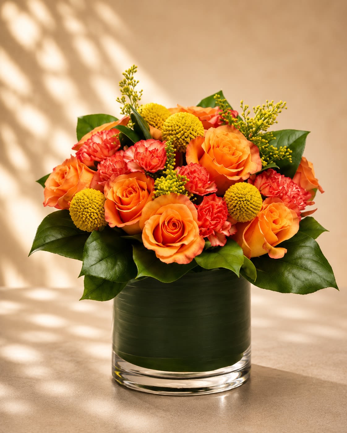 Golden Citrus Charm - A radiant and cheerful floral arrangement featuring glowing orange roses, vibrant coral carnations, playful yellow billy balls, and lush modern greenery in a sleek leaf-lined glass vase. Golden Citrus Charm blends warm citrus tones with rich textures for a design that feels fresh, uplifting, and effortlessly luxurious.  Orange roses symbolize enthusiasm, admiration, and joyful energy, while coral carnations add warmth and affection. The whimsical billy balls bring a contemporary designer touch that makes this arrangement feel artistic and unique. Styled with glossy greens and airy golden accents, this arrangement is perfect for birthdays, congratulations, celebrations, thank-you gifts, or simply brightening someone’s day.  Due to seasonal and market availability, specific accent flowers and greenery may vary slightly while maintaining the arrangement’s signature vibrant citrus-inspired palette and modern upscale style. Vase style may occasionally vary depending on availability while preserving the same elegant presentation.