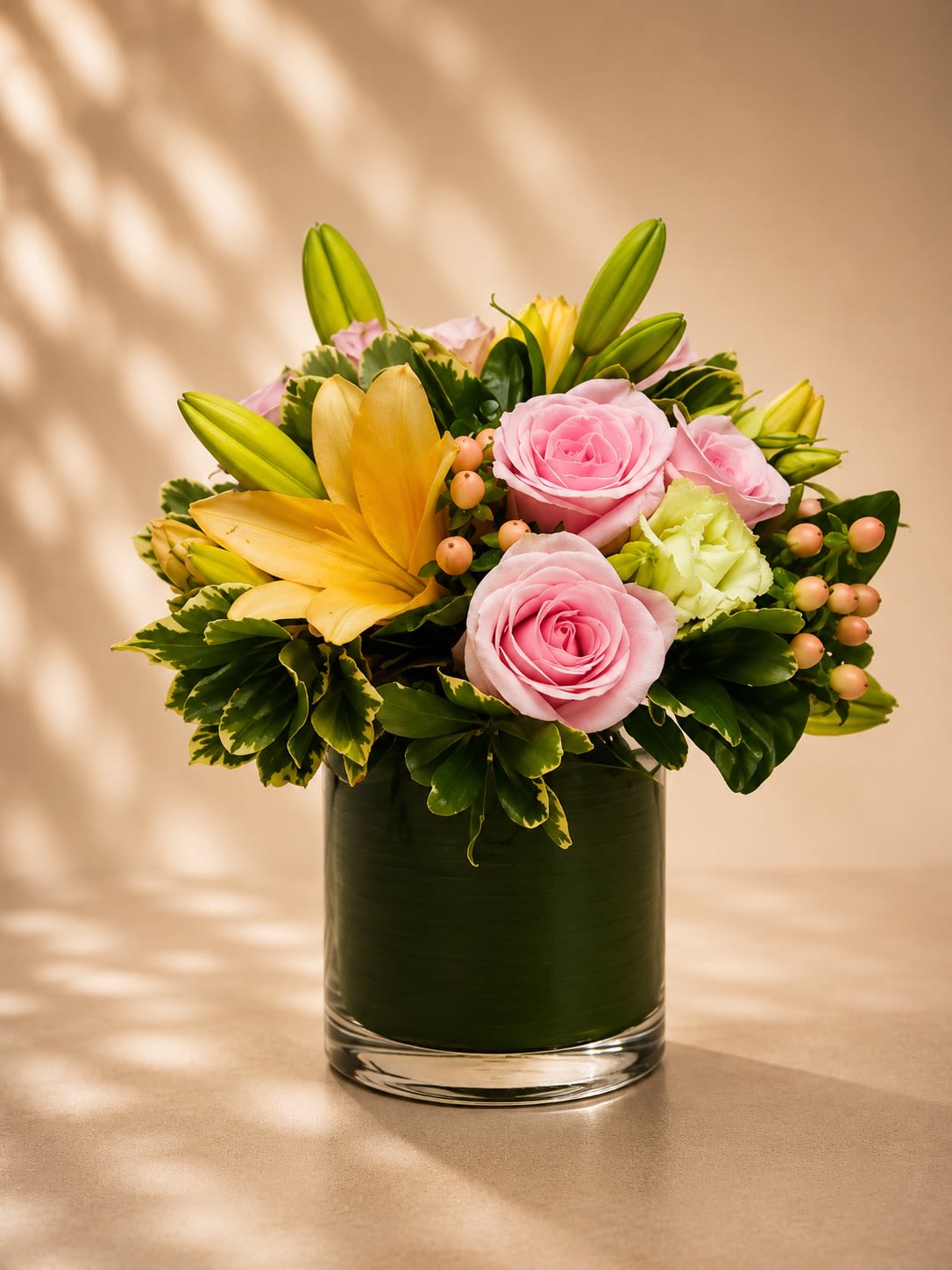 Blush Soleil Garden - A soft and radiant floral arrangement featuring delicate pink roses, golden lilies, fresh green accents, and blush hypericum berries arranged in a sleek leaf-lined glass vase. Blush Soleil Garden blends romantic pastel tones with warm golden hues for a design that feels graceful, cheerful, and luxuriously modern.  Pink roses symbolize admiration, sweetness, and elegance, while golden lilies represent happiness, positivity, and abundance. Accented with lush variegated greenery and soft berry textures, this arrangement creates a balanced designer look that feels both refined and welcoming. Perfect for birthdays, baby showers, thank-you gifts, celebrations, get well wishes, or simply brightening someone’s day with elegance and warmth.  Due to seasonal and market availability, lilies may arrive in various blooming stages to ensure longer-lasting beauty. Accent flowers and greenery may vary slightly while maintaining the arrangement’s signature soft romantic palette and upscale garden-inspired style. Vase style may occasionally vary depending on availability while preserving the same elegant presentation.