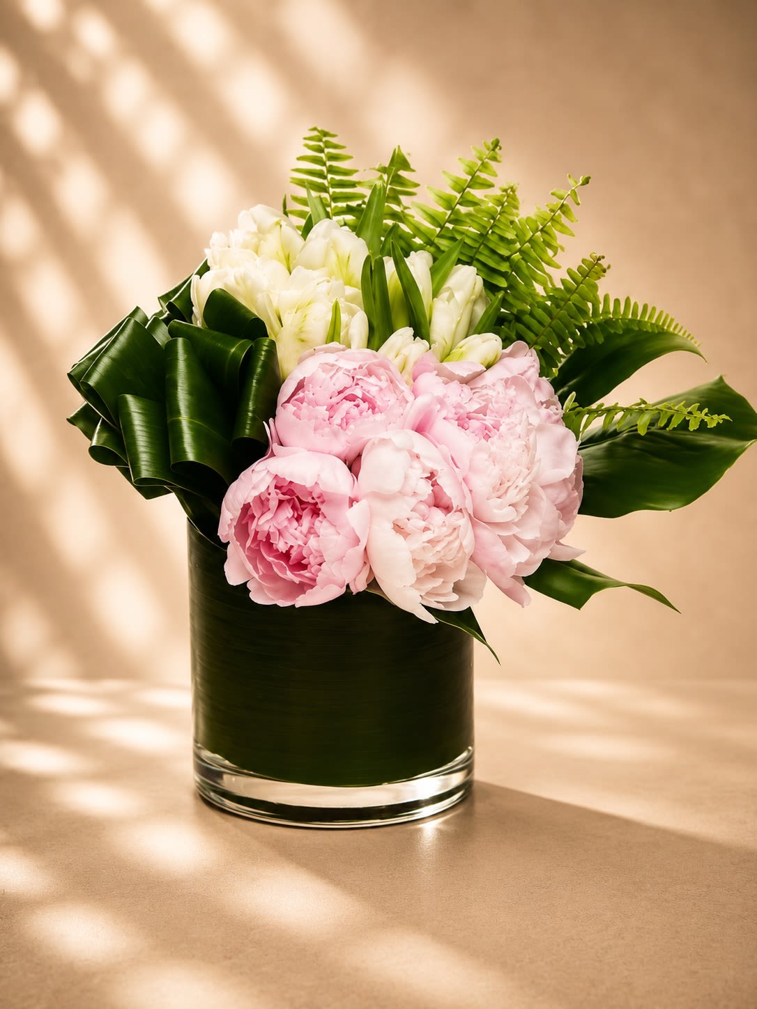 Blush Jardin Luxe - A refined garden-style arrangement featuring lush blush pink peonies, crisp white tulips, sculptural tropical foliage, and fresh fern accents designed in a sleek leaf-lined glass vase. Blush Jardin Luxe blends romantic softness with modern botanical elegance, creating a floral statement that feels serene, sophisticated, and effortlessly luxurious.  Peonies symbolize romance, prosperity, and beauty, while white tulips represent grace, peace, and new beginnings. The dramatic folded tropical leaves and airy fern textures add a contemporary designer touch, giving this arrangement a fresh editorial-inspired feel. Perfect for anniversaries, luxury gifting, birthdays, baby showers, sympathy sentiments, or elegant home décor.  Due to seasonal and market availability, peonies may occasionally be substituted with premium garden roses or similar luxury blooms while preserving the arrangement’s signature soft blush palette and upscale botanical design. Tulips may arrive in varying bloom stages for longer-lasting beauty. Vase style and greenery may vary slightly while maintaining the same elegant presentation.