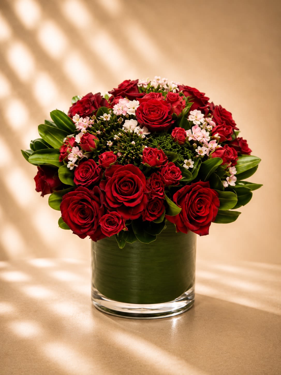 Ruby Love Garden - Elegant, romantic, and timeless, Ruby Love Garden is a lush arrangement of premium red roses and delicate spray roses accented with soft blush wax flowers and rich layered greenery in a modern leaf-lined glass vase.  Designed with a luxurious garden-inspired style, this arrangement blends classic romance with contemporary sophistication. The deep ruby roses symbolize passionate love, devotion, and elegance, while the miniature spray roses and delicate floral accents add softness, texture, and charm.  Its rounded silhouette creates a full abundant presentation that feels luxurious without being overwhelming — perfect for anniversaries, romantic celebrations, birthdays, Valentine’s Day, or upscale everyday gifting.  The contrast of velvety red blooms against glossy green foliage gives this arrangement a couture floral look inspired by high-end boutique florals and modern luxury design.