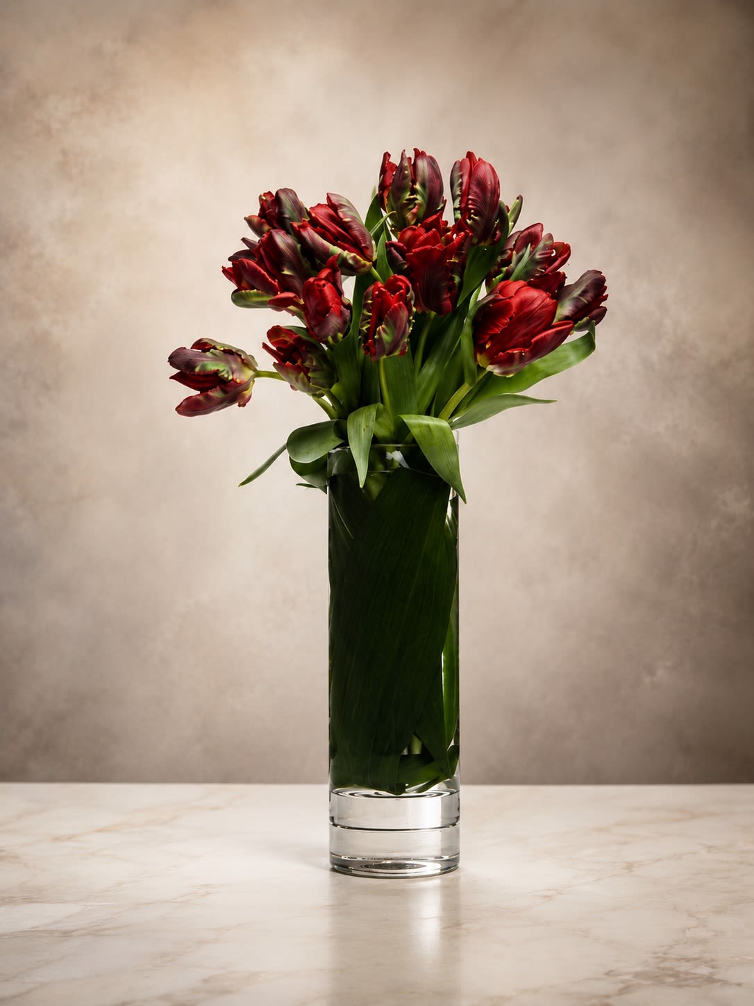 Midnight Tulips - Moody, dramatic, and undeniably sophisticated, Midnight Velvet Tulips features luxurious deep burgundy tulips arranged in a sleek tall cylinder vase wrapped with rich tropical foliage for a refined contemporary presentation.  The velvety wine-colored tulips create a bold statement of elegance and mystery, symbolizing deep passion, luxury, strength, and timeless beauty. Their sculptural shape and rich tones give this arrangement a modern editorial feel inspired by upscale European floral design.  Minimal yet striking, this arrangement is perfect for luxury gifting, romantic occasions, modern home décor, corporate settings, sympathy gestures, or customers seeking something unique and fashion-forward.  The tall clean silhouette paired with dramatic monochromatic blooms creates a high-end artistic aesthetic that feels both chic and unforgettable.