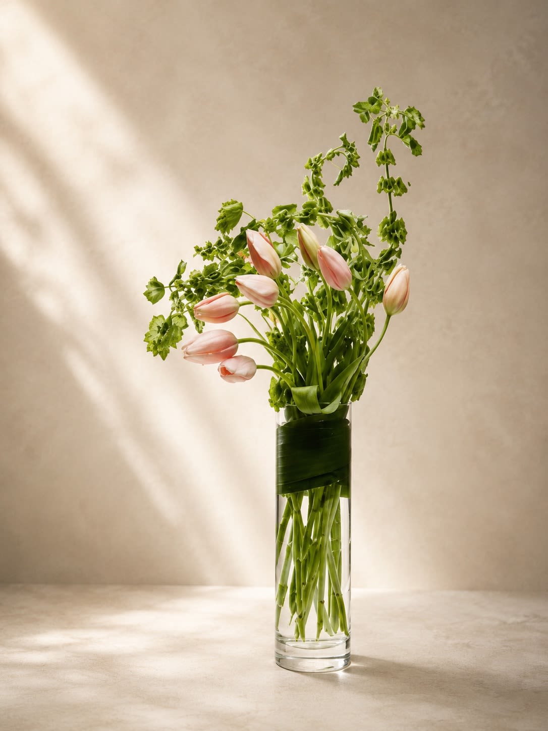 Blush Whisper Tulips - Soft, airy, and effortlessly artistic, Blush Whisper Tulips is a modern luxury arrangement designed with delicate blush pink tulips paired with flowing botanical greenery in a sleek clear cylinder vase wrapped with tropical leaf detailing.  This design embraces minimalist elegance with graceful movement and natural texture. The pale pink tulips symbolize gentle love, admiration, peace, and new beginnings, while the whimsical greenery adds a contemporary garden-inspired feel that feels fresh and refined.  Its tall sculptural silhouette creates a sophisticated statement perfect for luxury interiors, birthdays, sympathy gestures, baby showers, thank-you gifts, or simply elevating a modern space with understated beauty.  The clean lines, soft blush tones, and airy composition give this arrangement a high-end editorial aesthetic often seen in boutique hotels and luxury floral studios.