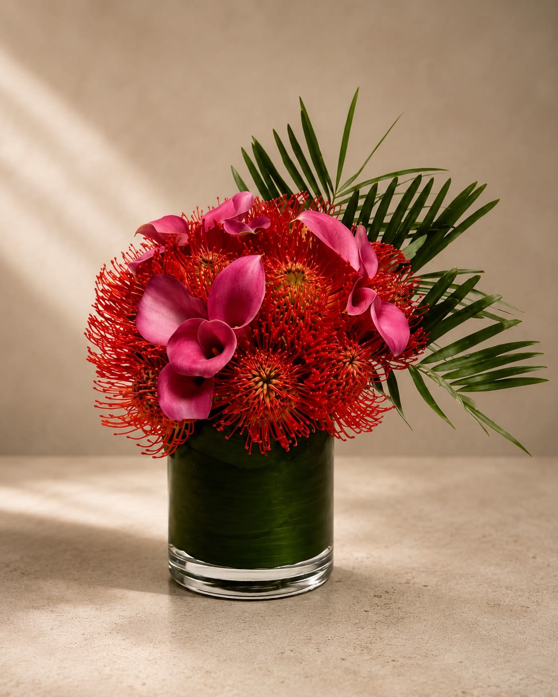 Crimson Tropique - Bold, artistic, and unapologetically luxurious, Crimson Tropique is a striking contemporary floral design inspired by exotic tropical elegance and high-fashion floral artistry.  This dramatic arrangement features vivid red pincushion protea paired with sculptural magenta calla lilies and sleek palm foliage, all arranged in a modern leaf-lined glass vase. The rich crimson and fuchsia tones create a daring, couture-inspired color palette that feels vibrant, passionate, and unforgettable.  The architectural textures and tropical elements give this arrangement a sophisticated editorial look often seen in luxury resorts, modern penthouses, and upscale designer events. Perfect for grand celebrations, luxury gifting, statement décor, birthdays, corporate spaces, or anyone who loves bold contemporary floral design.  The pincushion protea symbolizes strength, transformation, and creativity, while the calla lilies add elegance, beauty, and modern refinement.