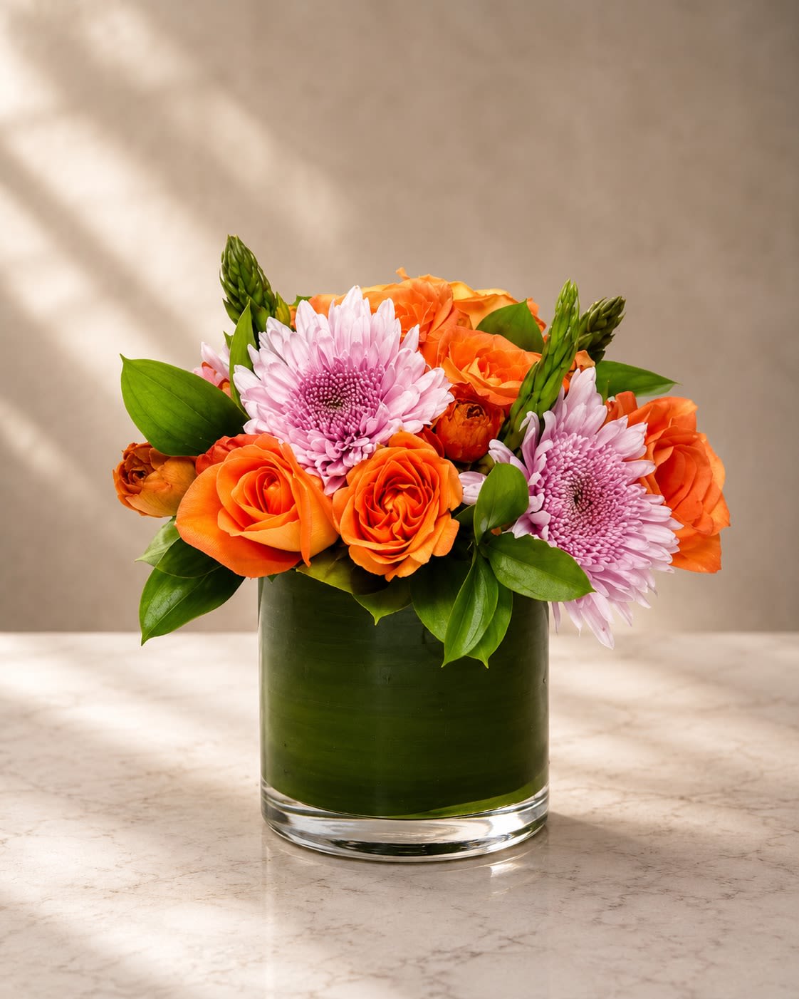 Sunset Blush Garden - Playful, cheerful, and beautifully refined, Sunset Blush Garden is a vibrant luxury arrangement inspired by warm golden sunsets and soft romantic tones.  This charming design features radiant orange roses paired with delicate blush spider mums and fresh seasonal greenery, all arranged in a sleek leaf-lined glass cylinder vase. The contrast between the vivid citrus-orange blooms and soft pastel pink florals creates a modern color palette that feels joyful, feminine, and effortlessly elegant.  The arrangement’s compact rounded shape gives it a polished boutique-style appearance while the layered textures add movement and sophistication. Perfect for birthdays, celebrations, thank-you gifts, spring occasions, friendship bouquets, or brightening any luxury interior space.  Orange roses symbolize enthusiasm, energy, and admiration, while the blush pink blooms bring softness, grace, and warmth to the design.