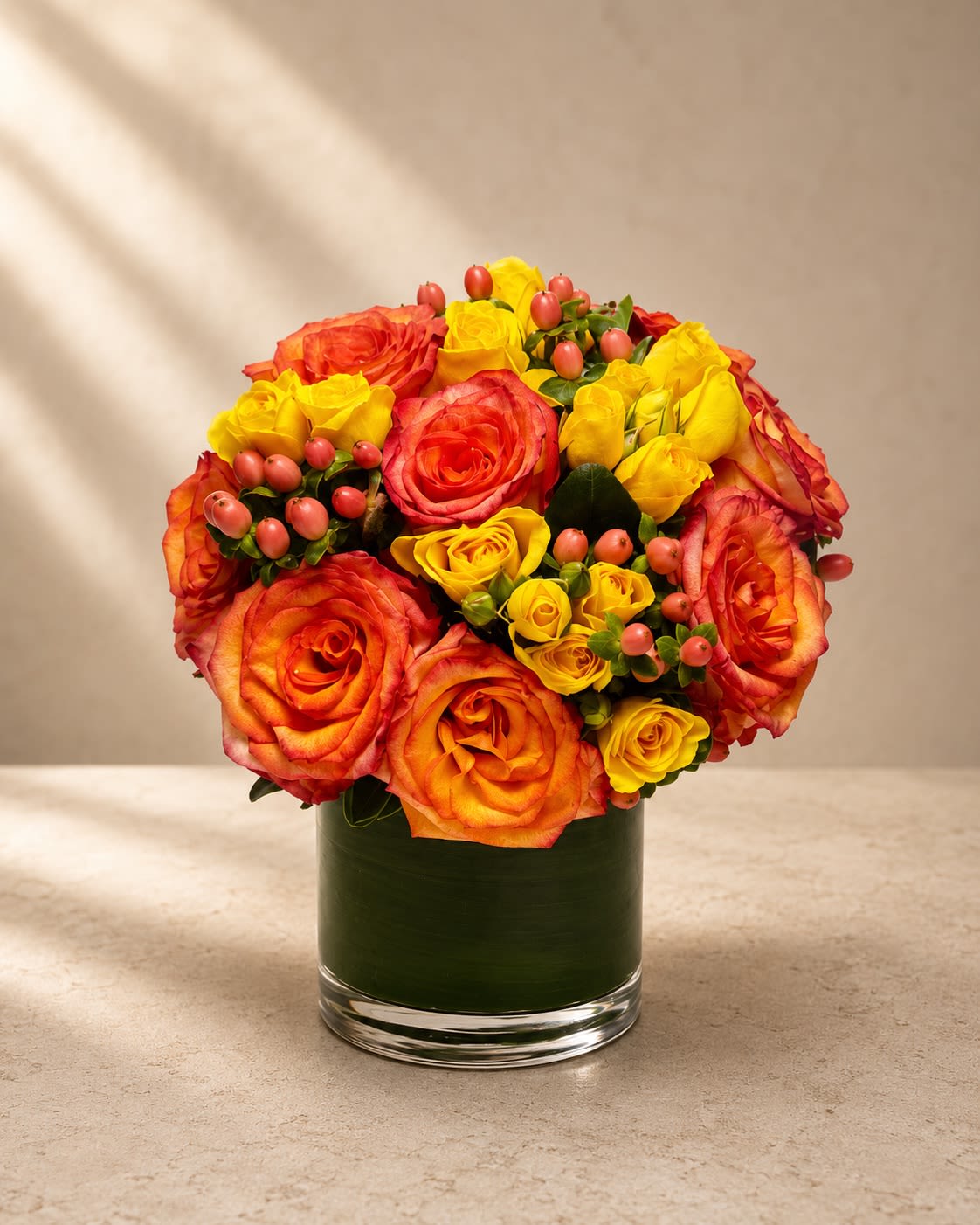 Citrus Ember Luxe - Vibrant, radiant, and full of modern energy, Citrus Ember Luxe is a bold luxury arrangement designed with glowing sunset-inspired tones and rich floral texture.  This striking design features premium orange roses paired with delicate yellow spray roses and vibrant coral hypericum berries, all artfully arranged in a sleek leaf-lined glass vase. The fiery blend of citrus orange, golden yellow, and warm coral creates a luminous palette that feels joyful, upscale, and fashion-forward.  The compact rounded silhouette gives the arrangement a polished boutique aesthetic while the layered rose varieties add depth and dimension. Perfect for birthdays, celebrations, congratulations, thank-you gifts, luxury hospitality décor, or anyone who loves vibrant contemporary floral design.  Orange roses symbolize enthusiasm, fascination, and confidence, while yellow roses bring warmth, happiness, and friendship — together creating a design that feels uplifting, luxurious, and unforgettable.